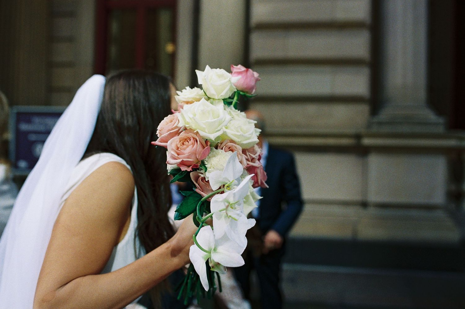 Bride in white dress and veil holding a cascading bouquet of pink and white roses and orchids outside a building.