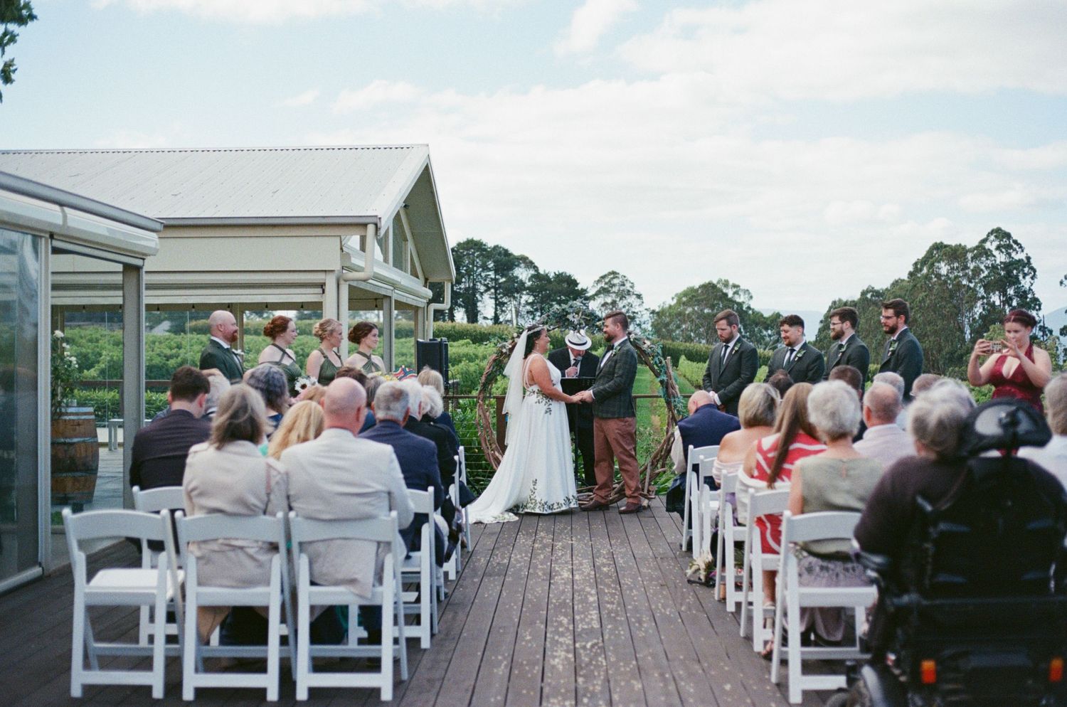 Outdoor wedding ceremony on a wooden deck with guests seated in white chairs, bride and groom exchanging vows.
