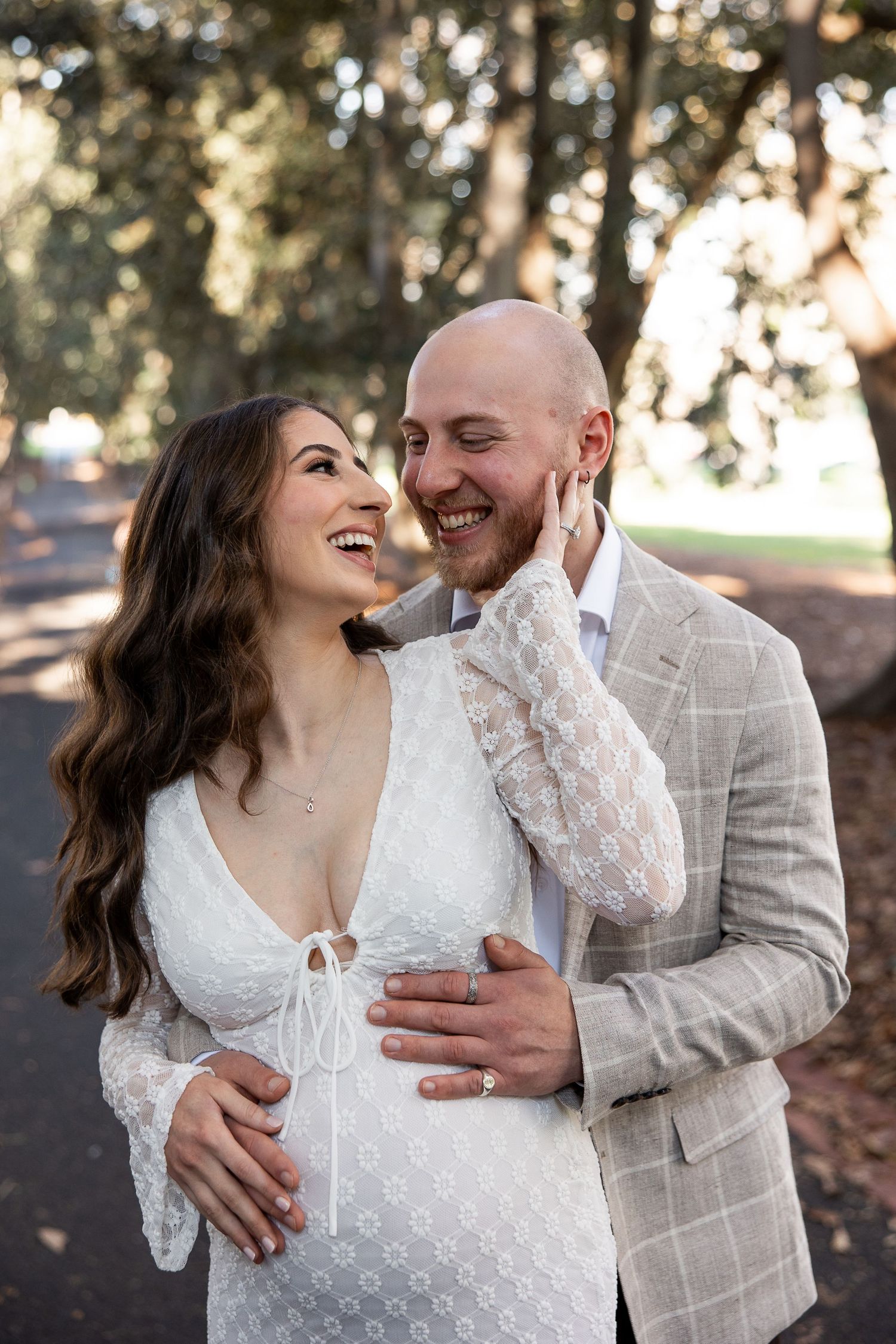 A romantic couple shares joyful laughter while embracing outdoors in a rustic setting during a wedding photoshoot.