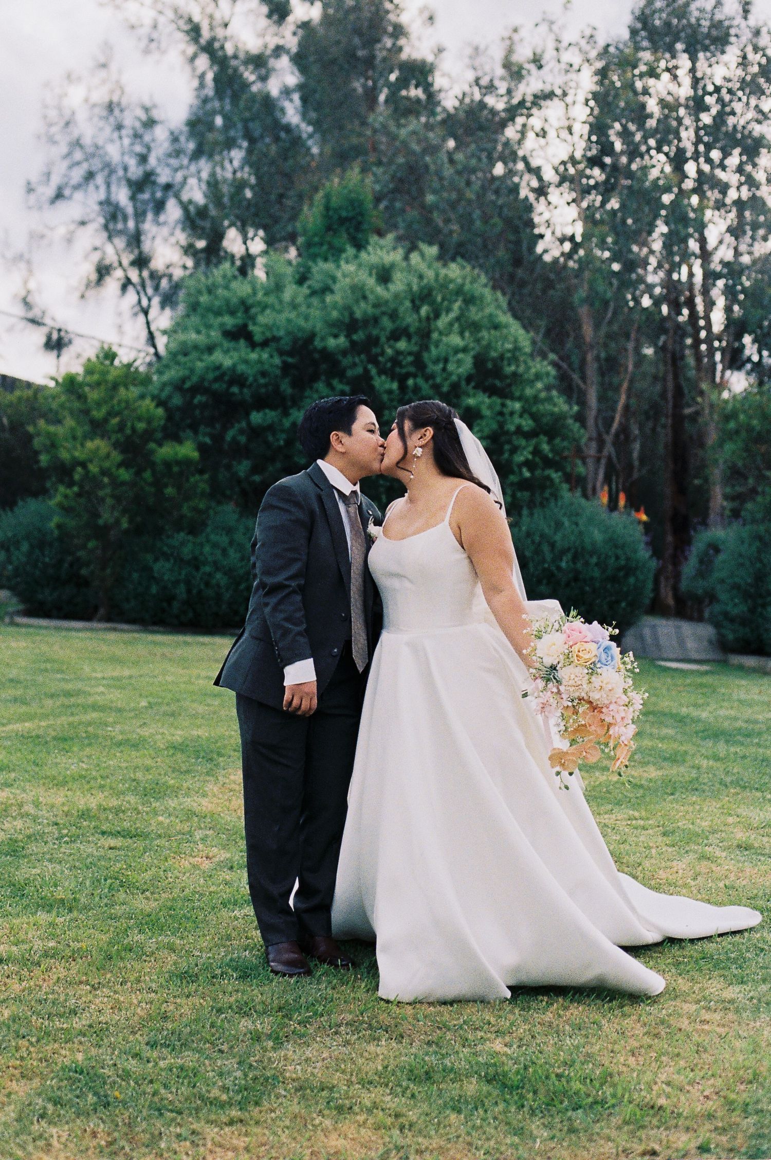 Bride and groom share a kiss on green lawn, she holds pastel bouquet, trees in background.