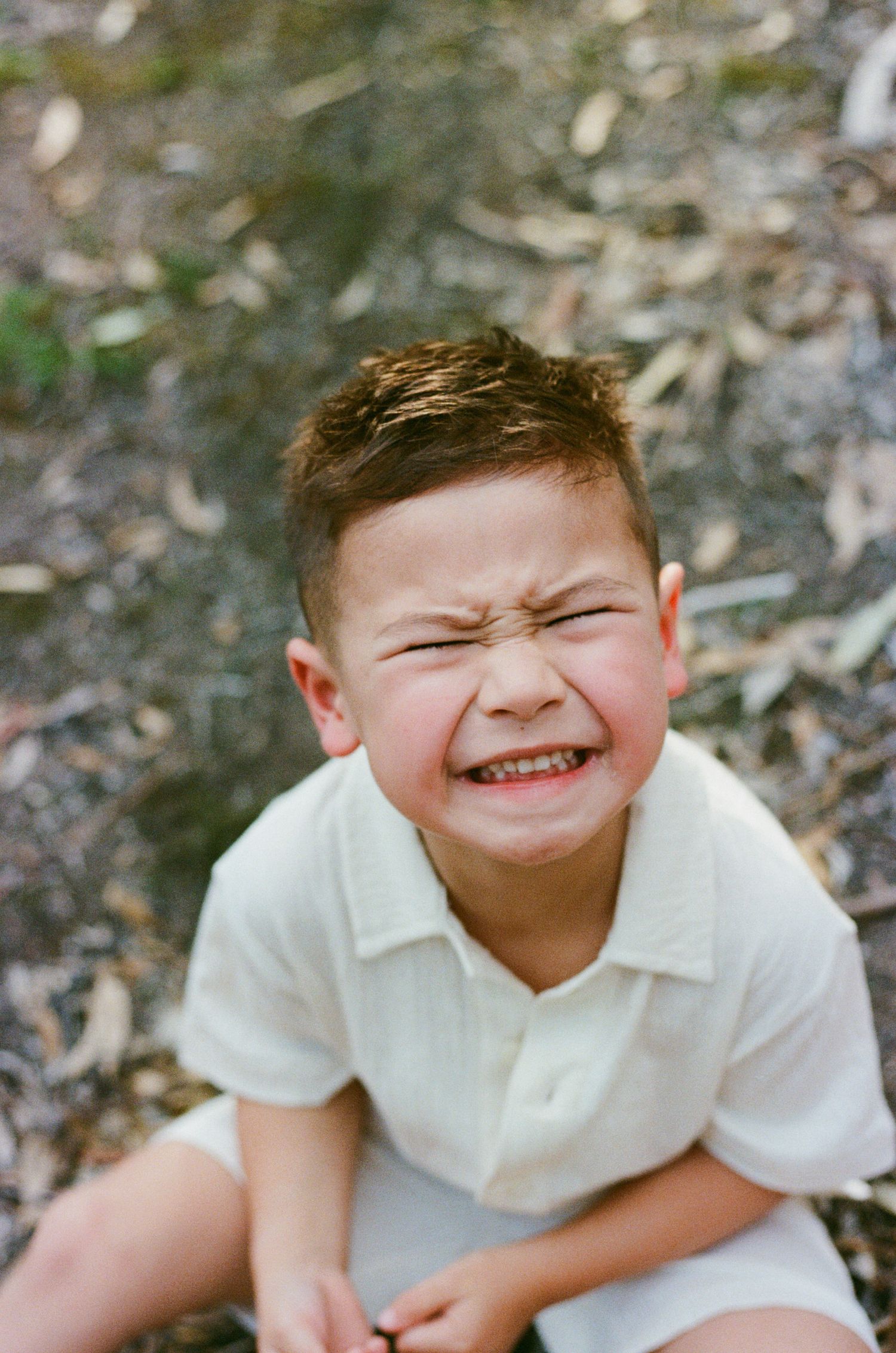 Young boy in white shirt making a funny scrunched face while sitting outdoors among fallen leaves.