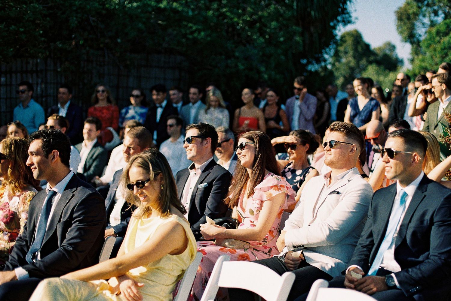 Guests in sunglasses and formal attire seated outdoors at a sunny wedding ceremony.