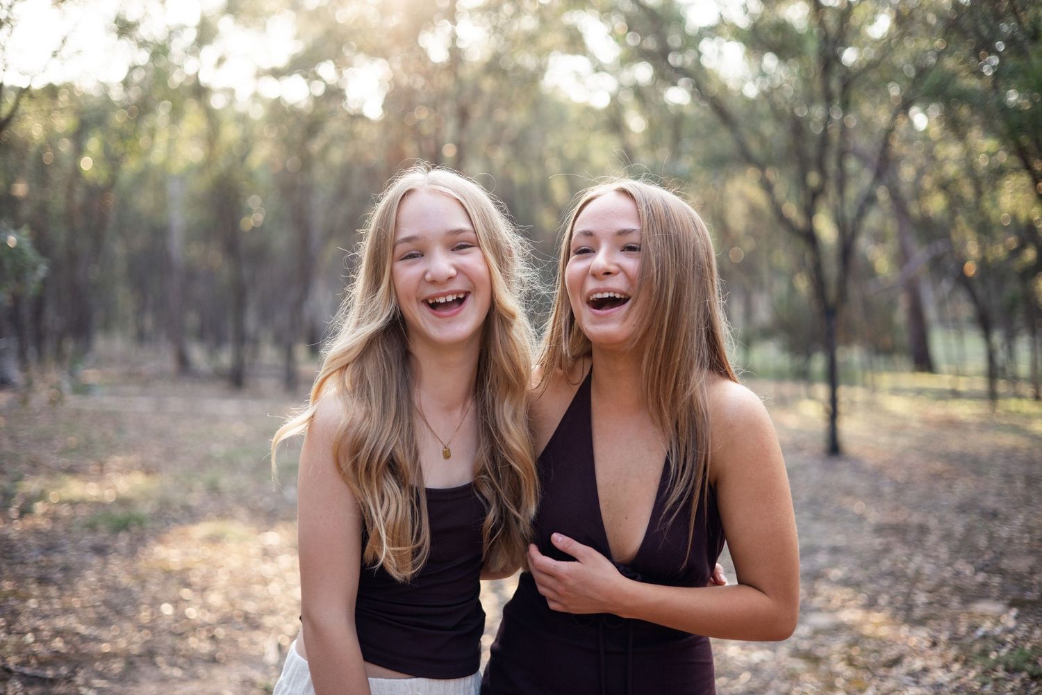 Two laughing teenage girls in black outfits pose together in a sunlit forest setting.