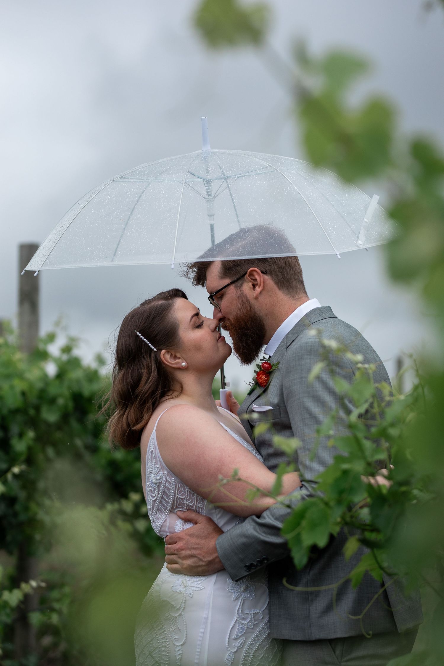 A romantic couple shares an intimate moment under a clear umbrella in a garden on a rainy day.