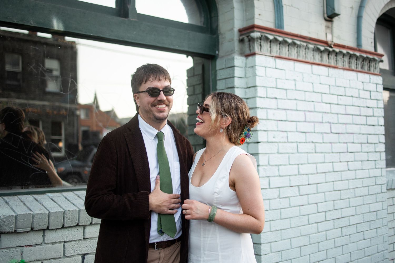 A couple smiles together outdoors, man in dark blazer and green tie, woman in white dress, against a white brick wall.