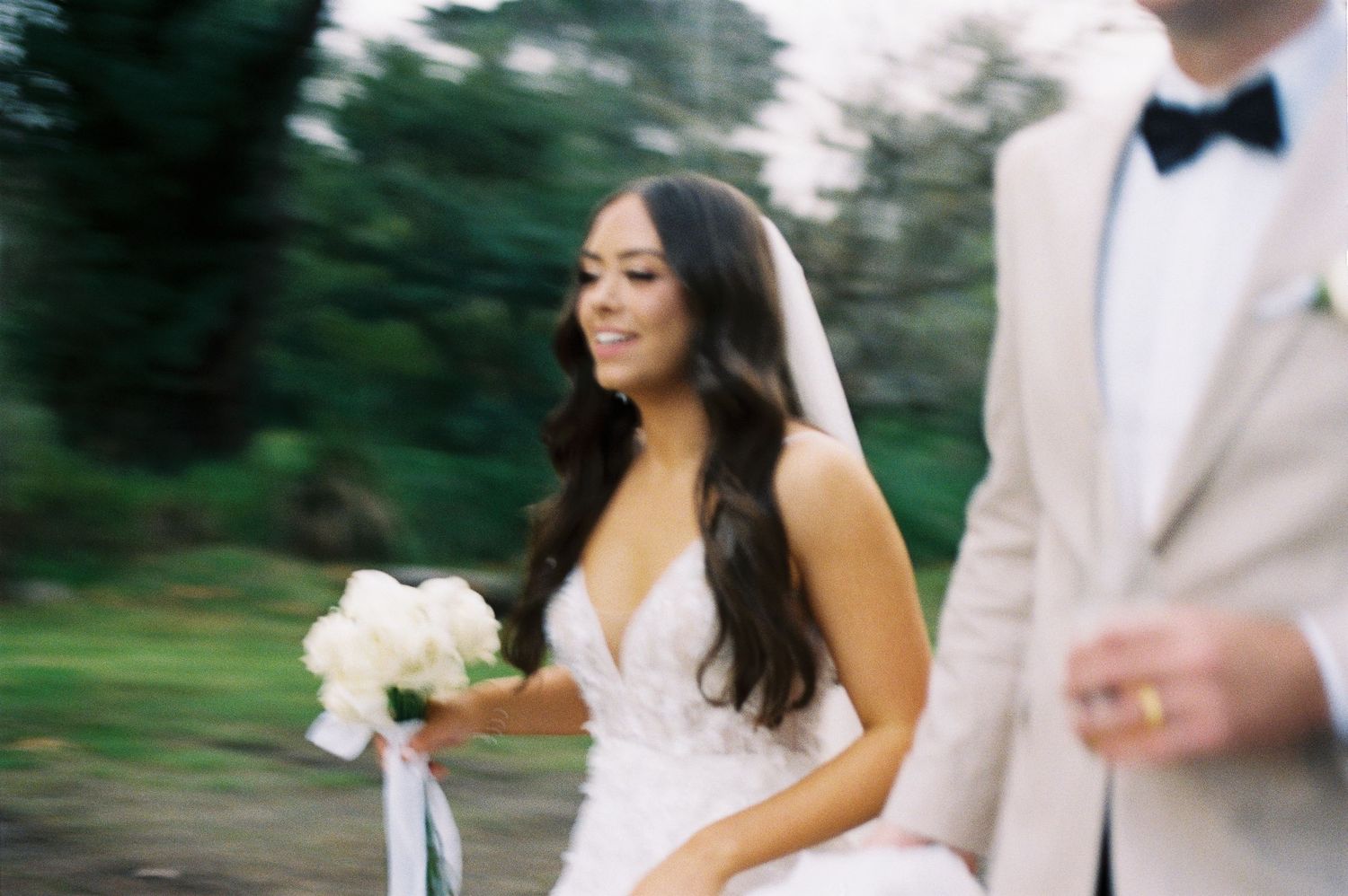 Bride in white gown holding bouquet walks beside groom in tan suit outdoors.