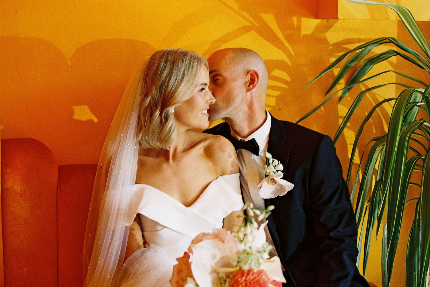 Bride and groom share a romantic kiss in front of a vibrant orange wall, bride holding a colorful floral bouquet.