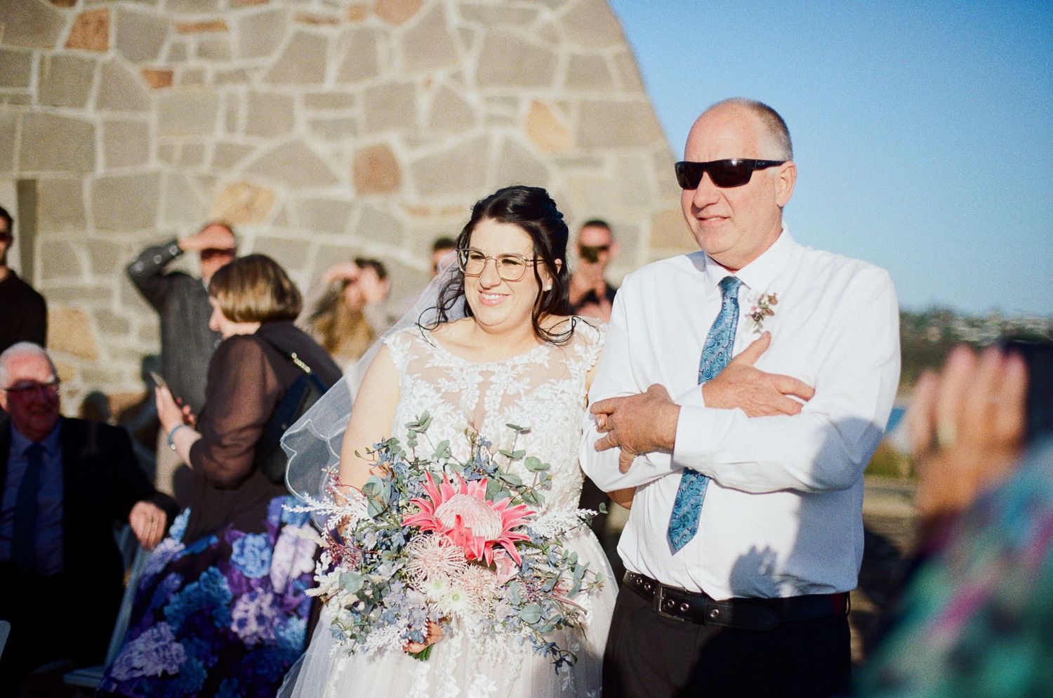 Wedding guests celebrate outdoors at a stone venue during a sunny afternoon with festive drinks and laughter.