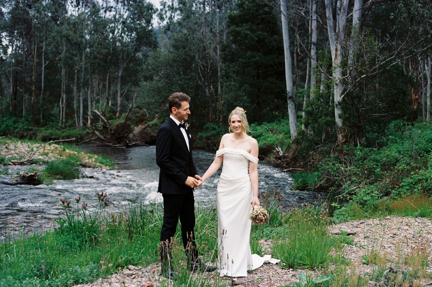 A couple in formal wedding attire walks together through a lush forest setting near a rocky stream.