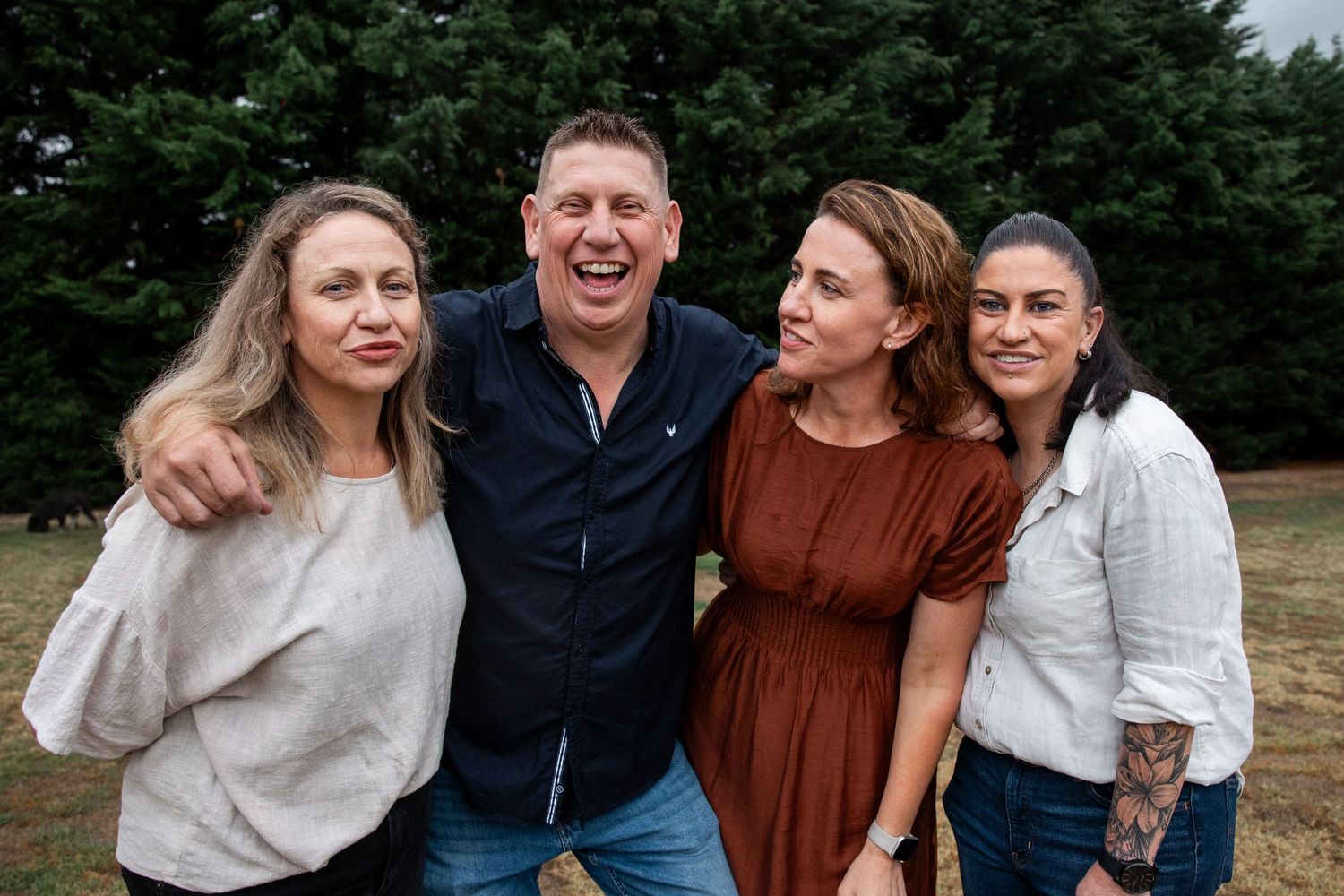 Four smiling adults posing together outdoors near green trees on a cloudy day.