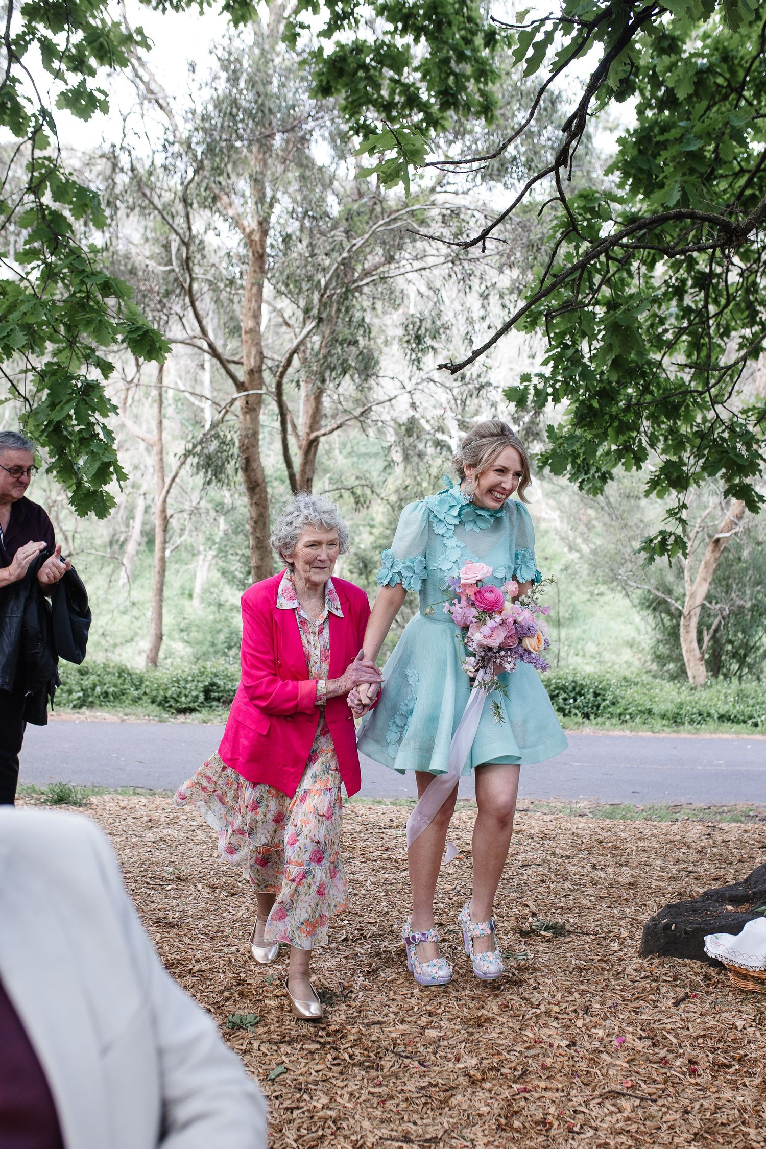 People dancing outdoors on woodchips surrounded by green trees and foliage in a park setting.