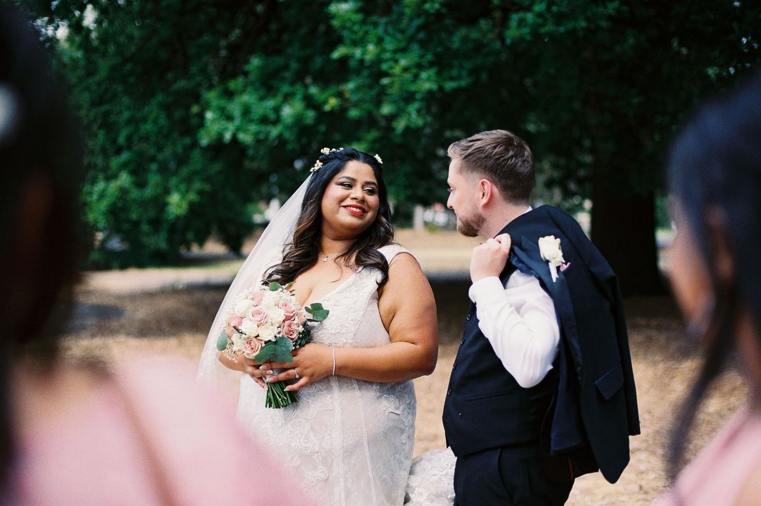 Bride and groom share a romantic moment outdoors, with the bride holding a pink bouquet in a lace wedding gown.