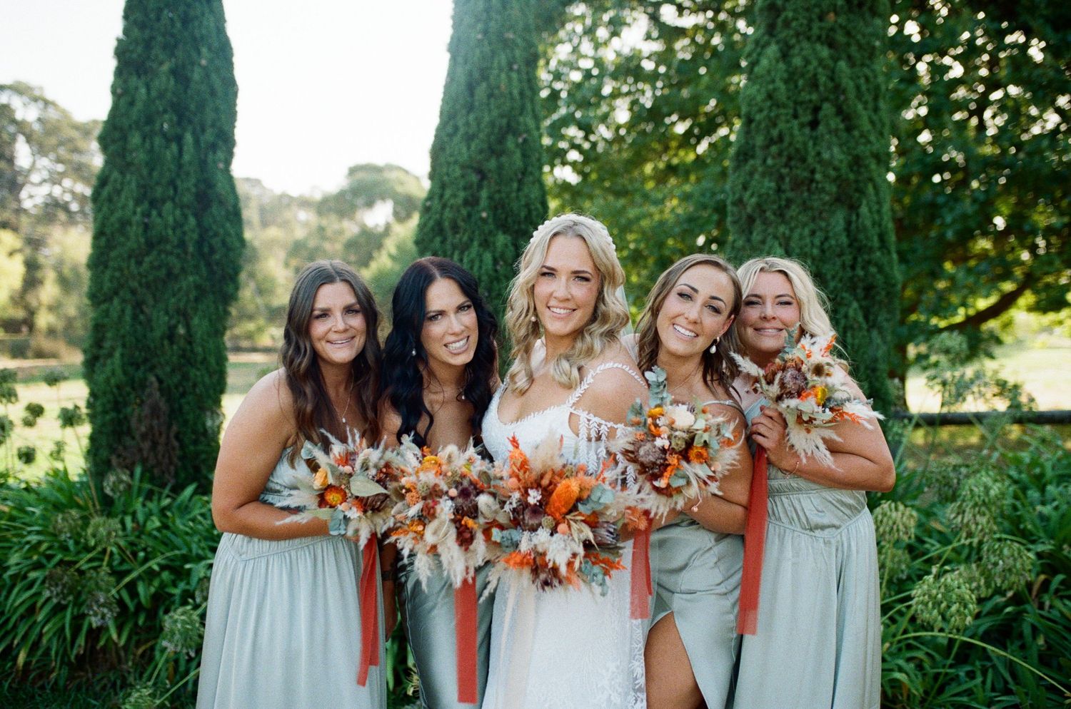 Bride in white gown with four bridesmaids in sage green dresses holding colorful bouquets outdoors.