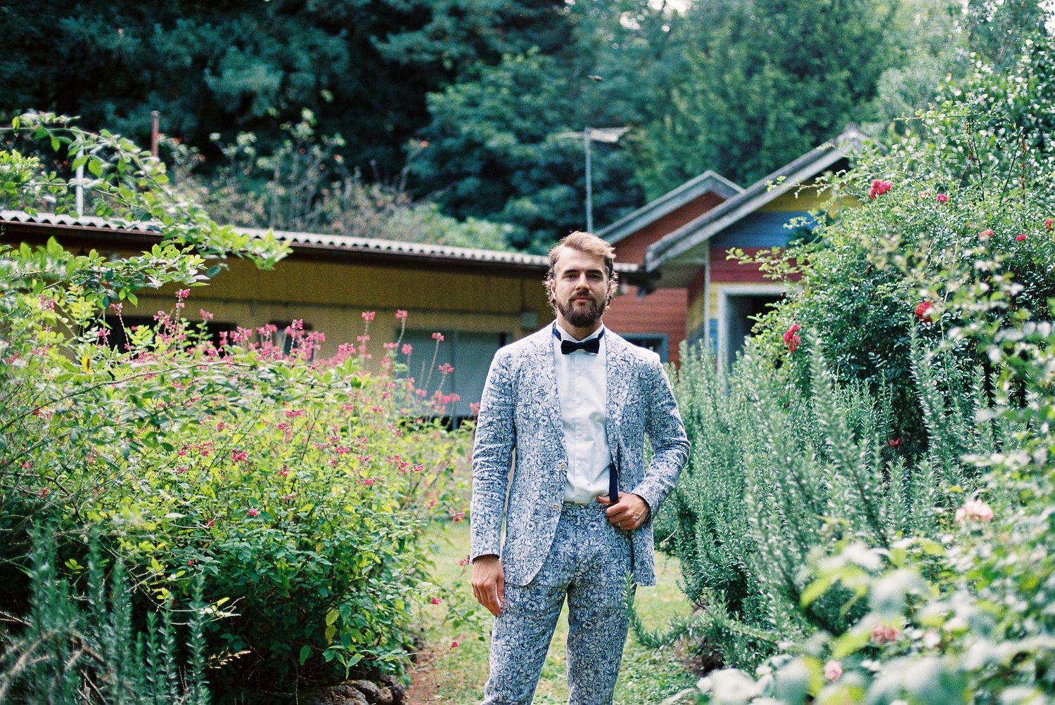 Man in patterned gray suit with bow tie standing in lush garden with colorful flowers and green foliage.