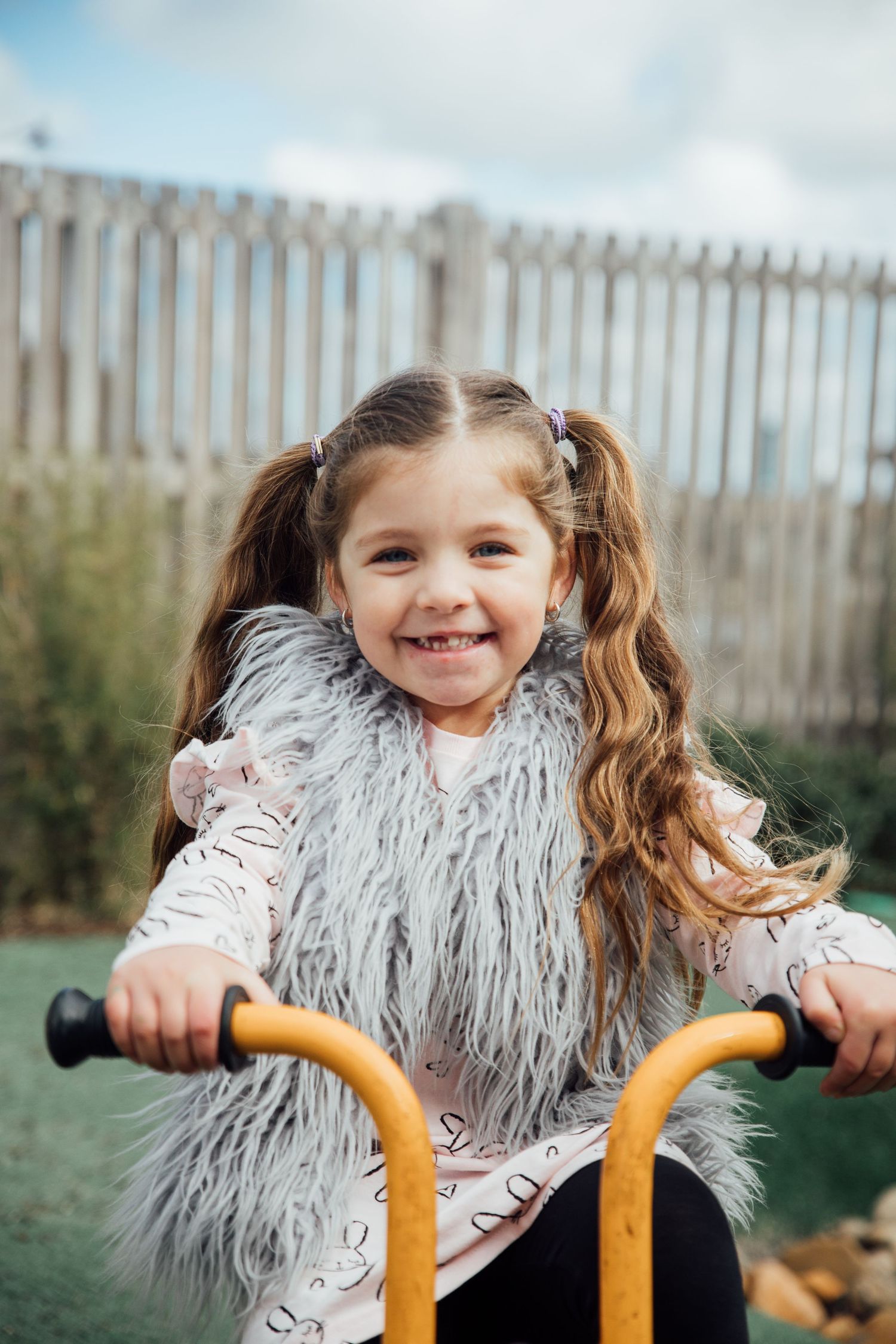 Small child in fluffy grey vest smiles while holding yellow handlebars of playground equipment outdoors.