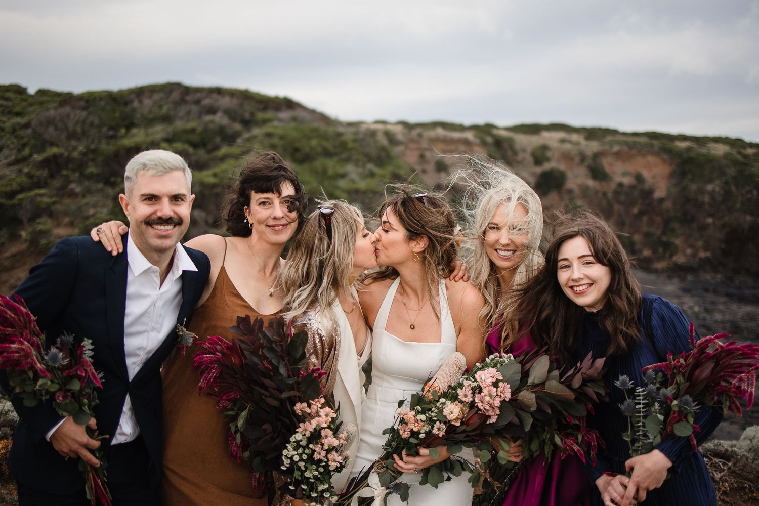 Wedding party gathered on coastal cliff with burgundy floral bouquets wearing rich jewel tones against dramatic landscape.