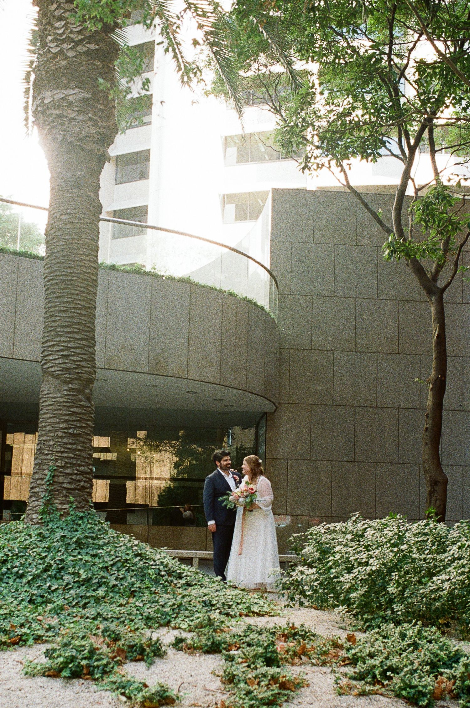 Bride and groom stand together in a lush courtyard beside a tall palm tree, with a modern stone building behind them.