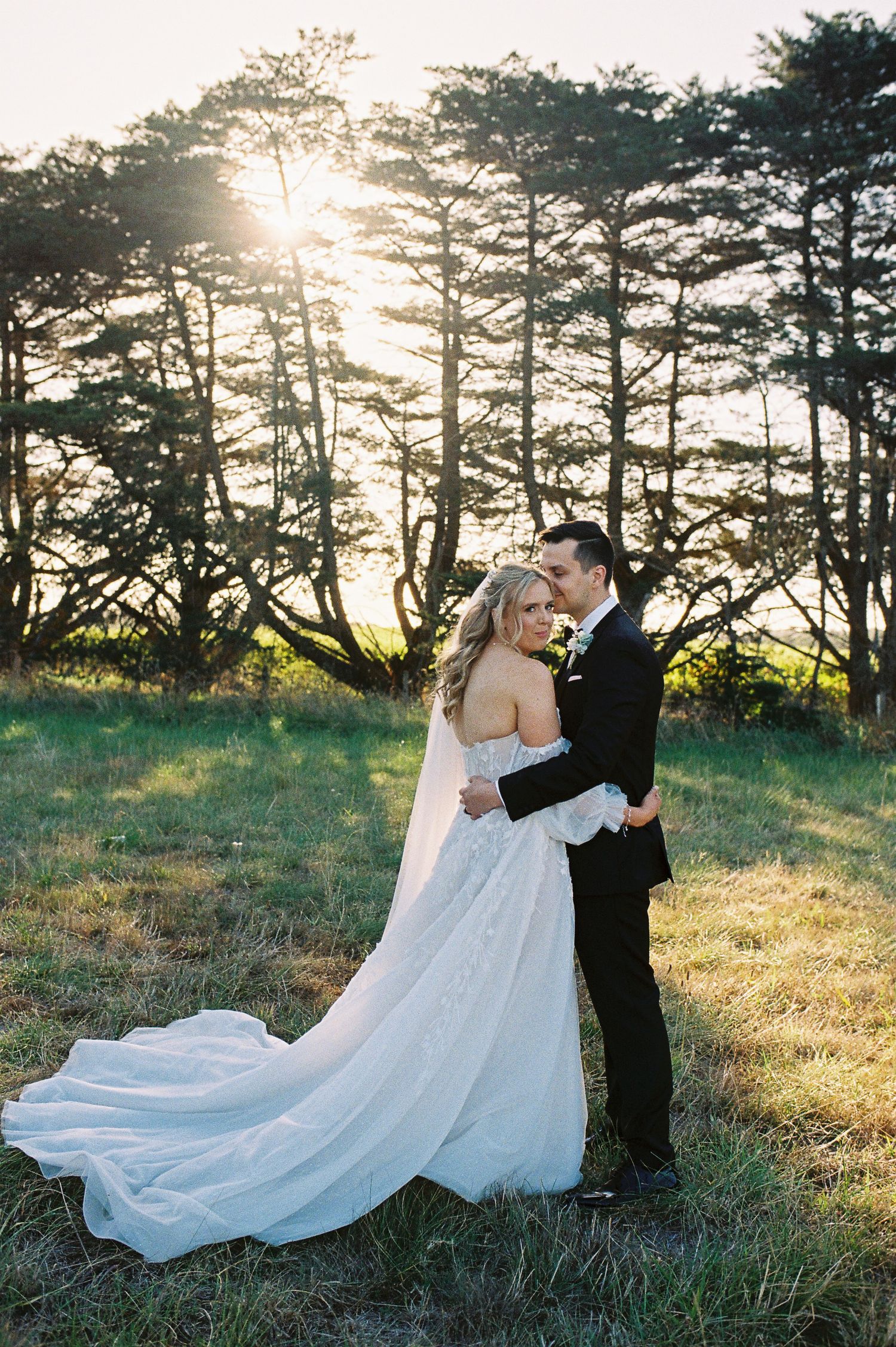 Bride and groom embrace at golden hour in a meadow, her lace gown trailing, sun rays glowing through tall trees behind them.