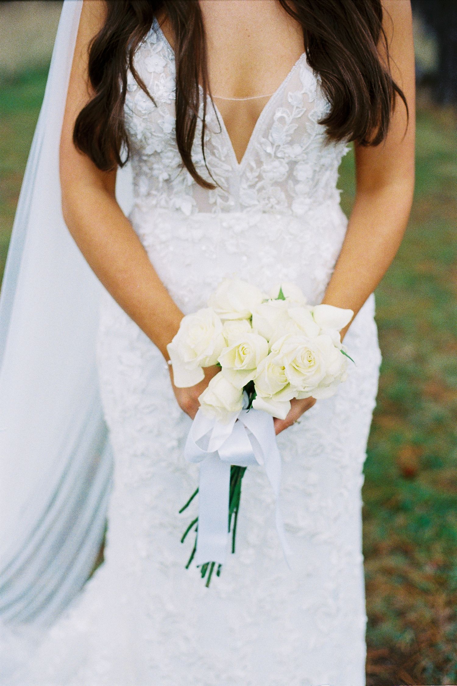Bride in lace wedding gown holding elegant white rose bouquet tied with ribbon.