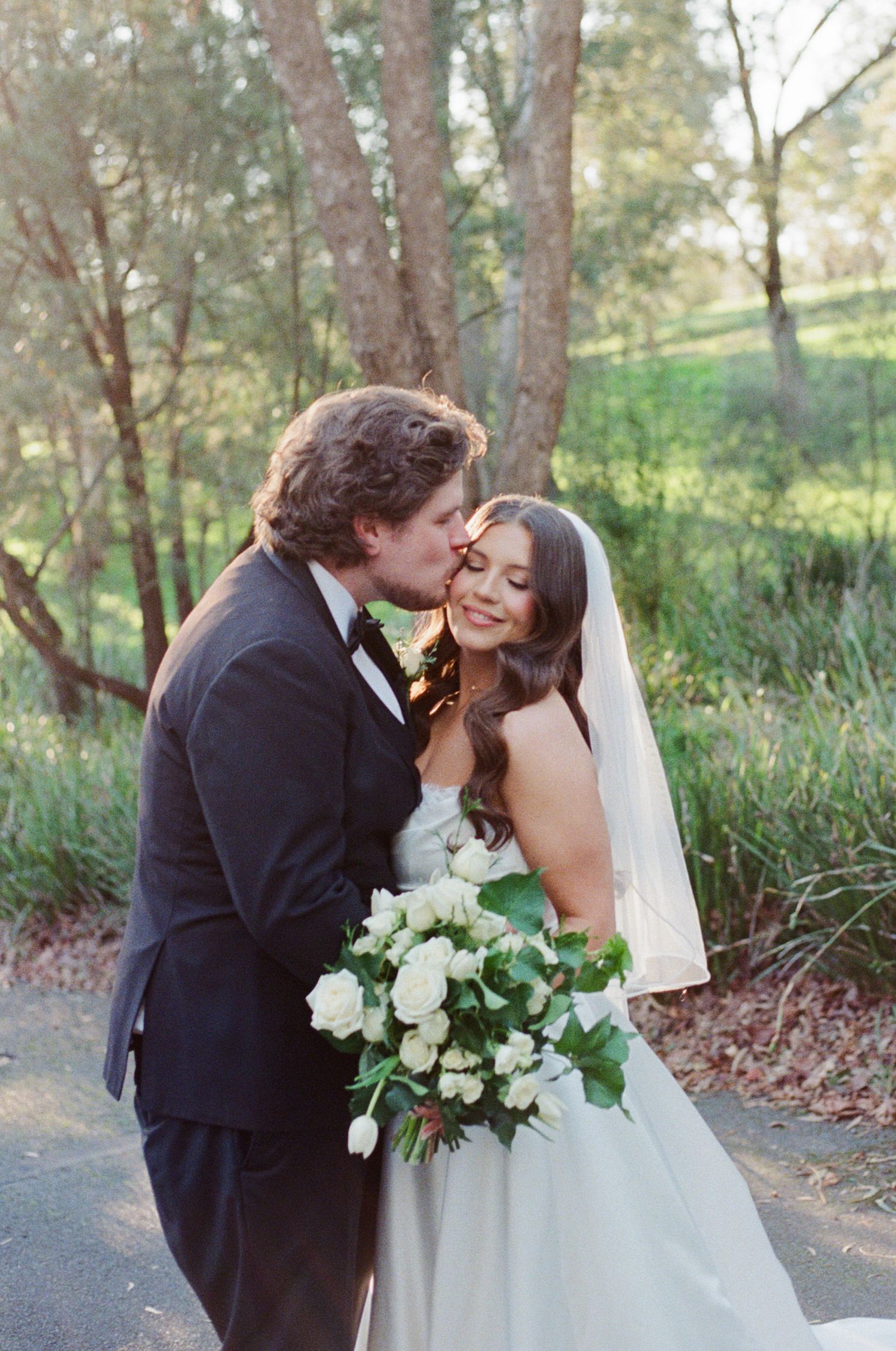 Groom in black tuxedo kisses bride wearing white gown and veil holding white floral bouquet outdoors.