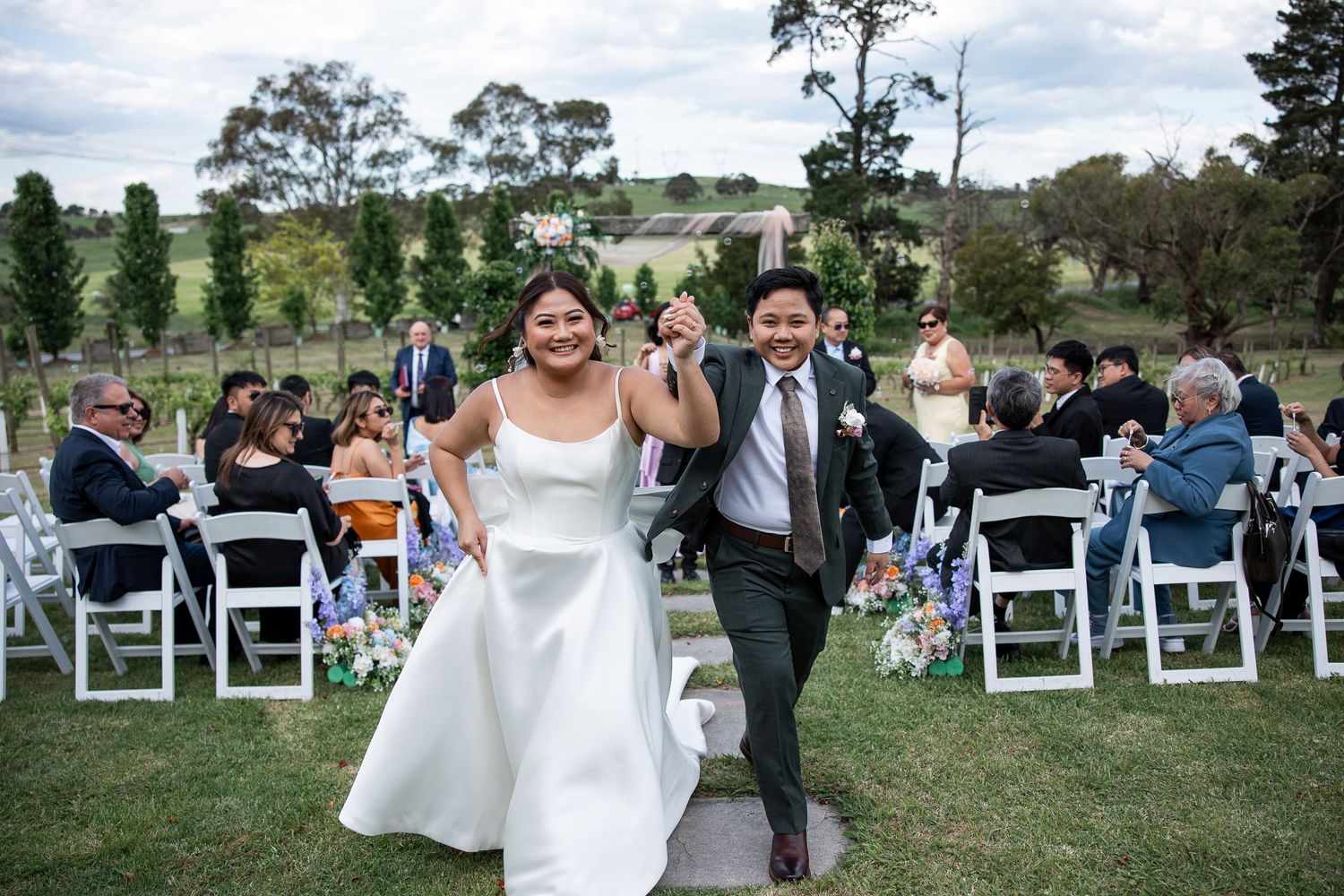 Happy couple exits their outdoor wedding ceremony on a grassy lawn with white chairs and guests celebrating behind them.