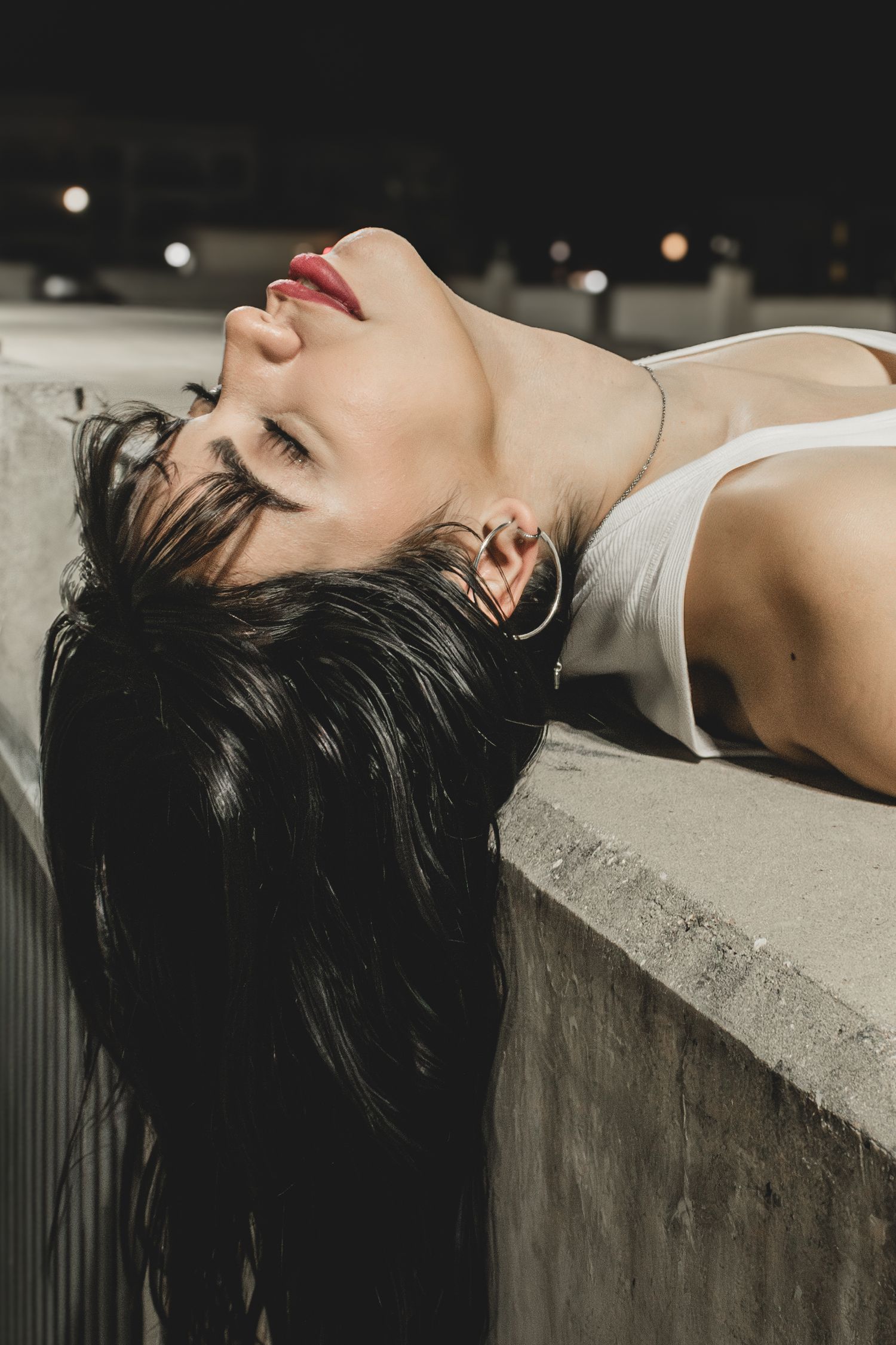 A person with dark hair and red lips lying back on a concrete ledge at night with bokeh lights in the background.