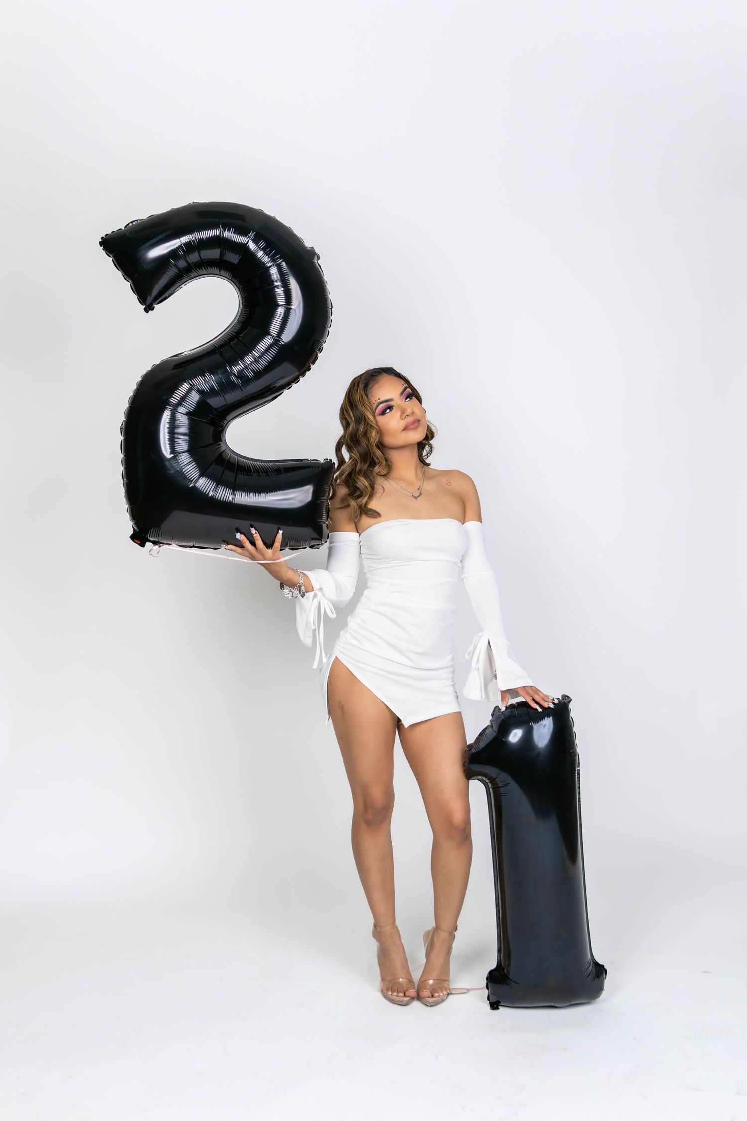 Person in white bodysuit poses with large black number 2 balloon against white background.