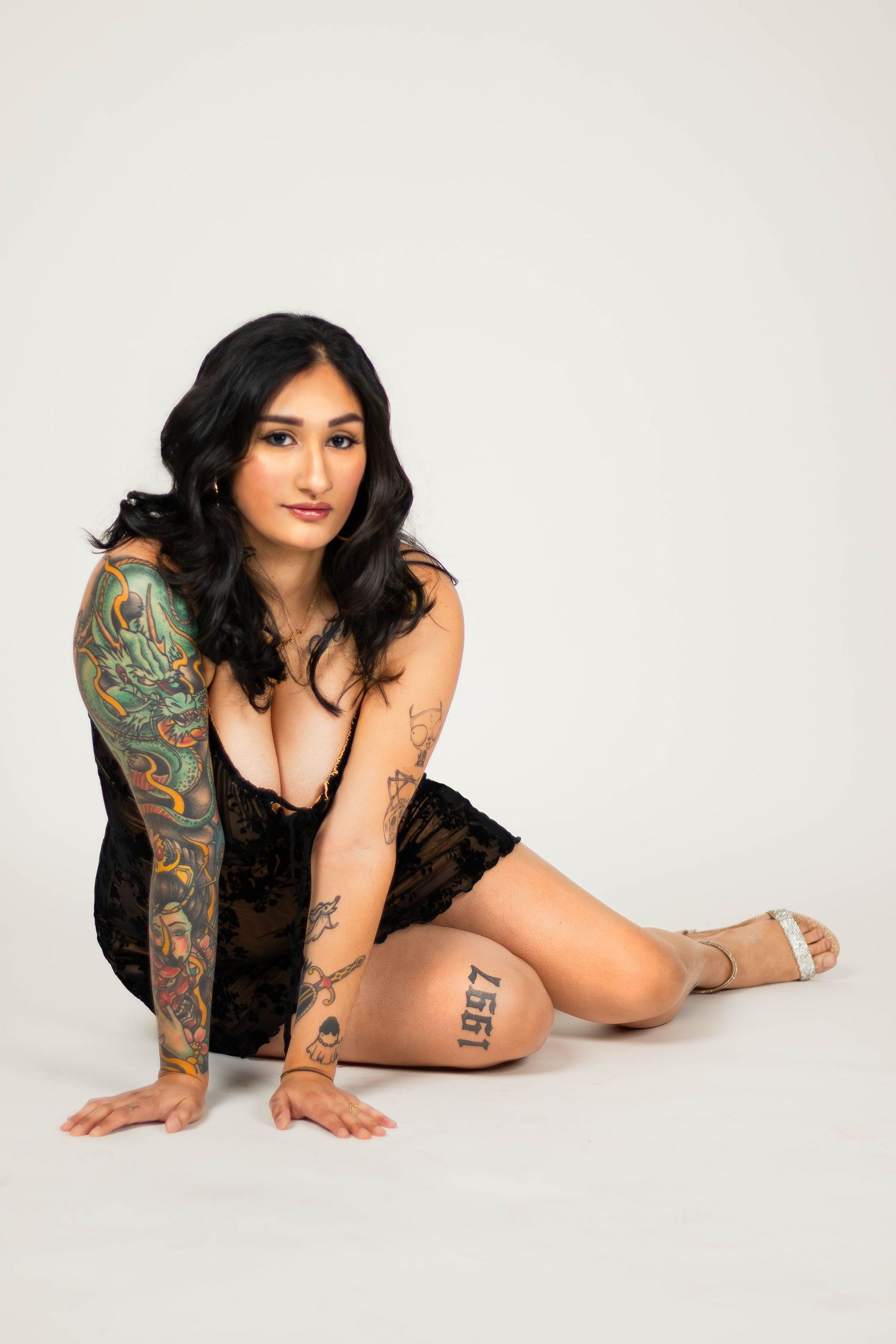 Model in black lingerie poses on white background showing colorful sleeve tattoo and smaller arm tattoos in San Antonio, Texas