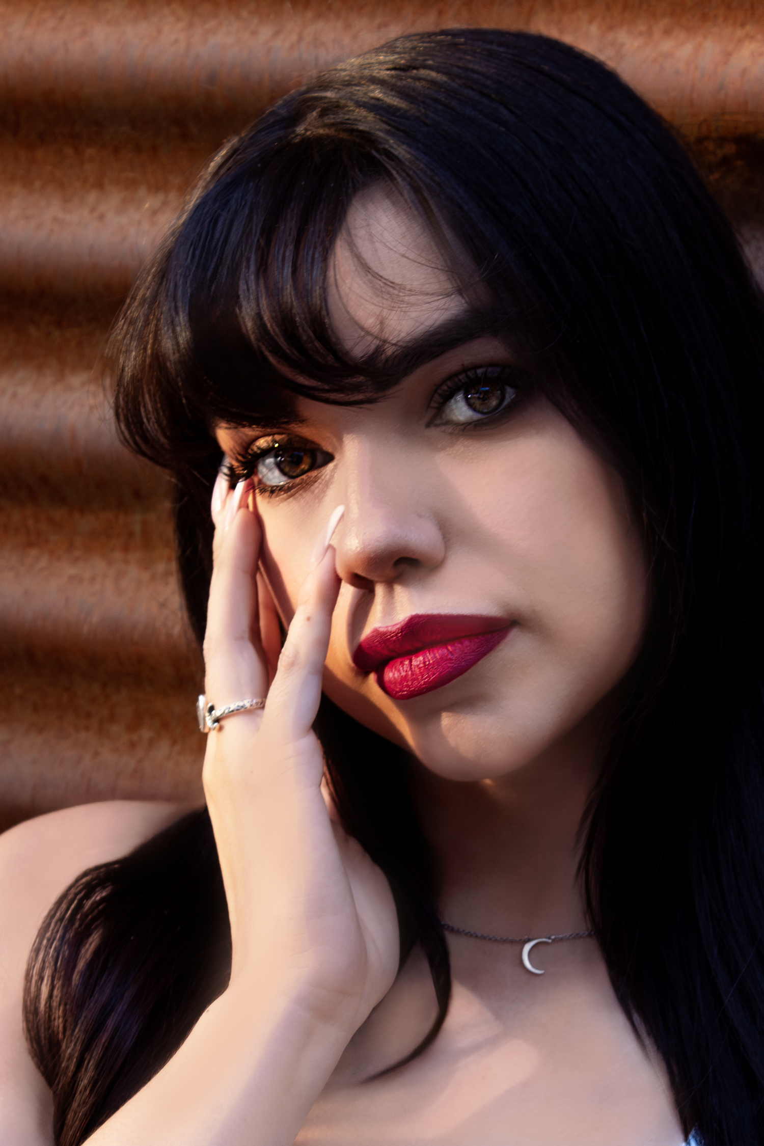 Portrait photo with bold red lipstick and dark bangs against a copper toned background wearing a crescent moon necklace.