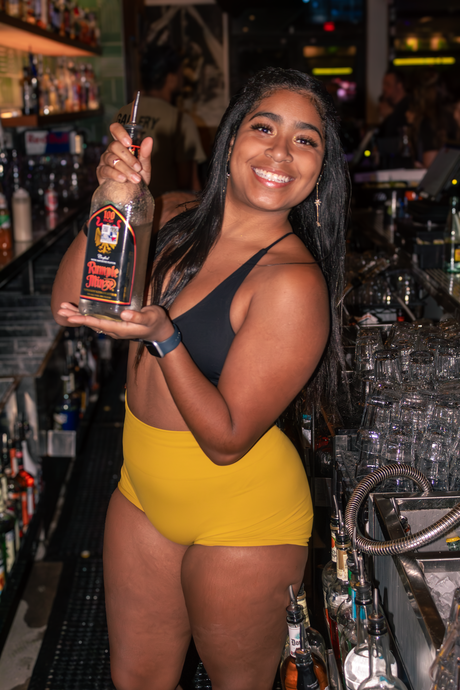 Bartender in black top and yellow shorts holds up a bottle while smiling at busy bar with glassware behind at Camp 1604 in San Antonio