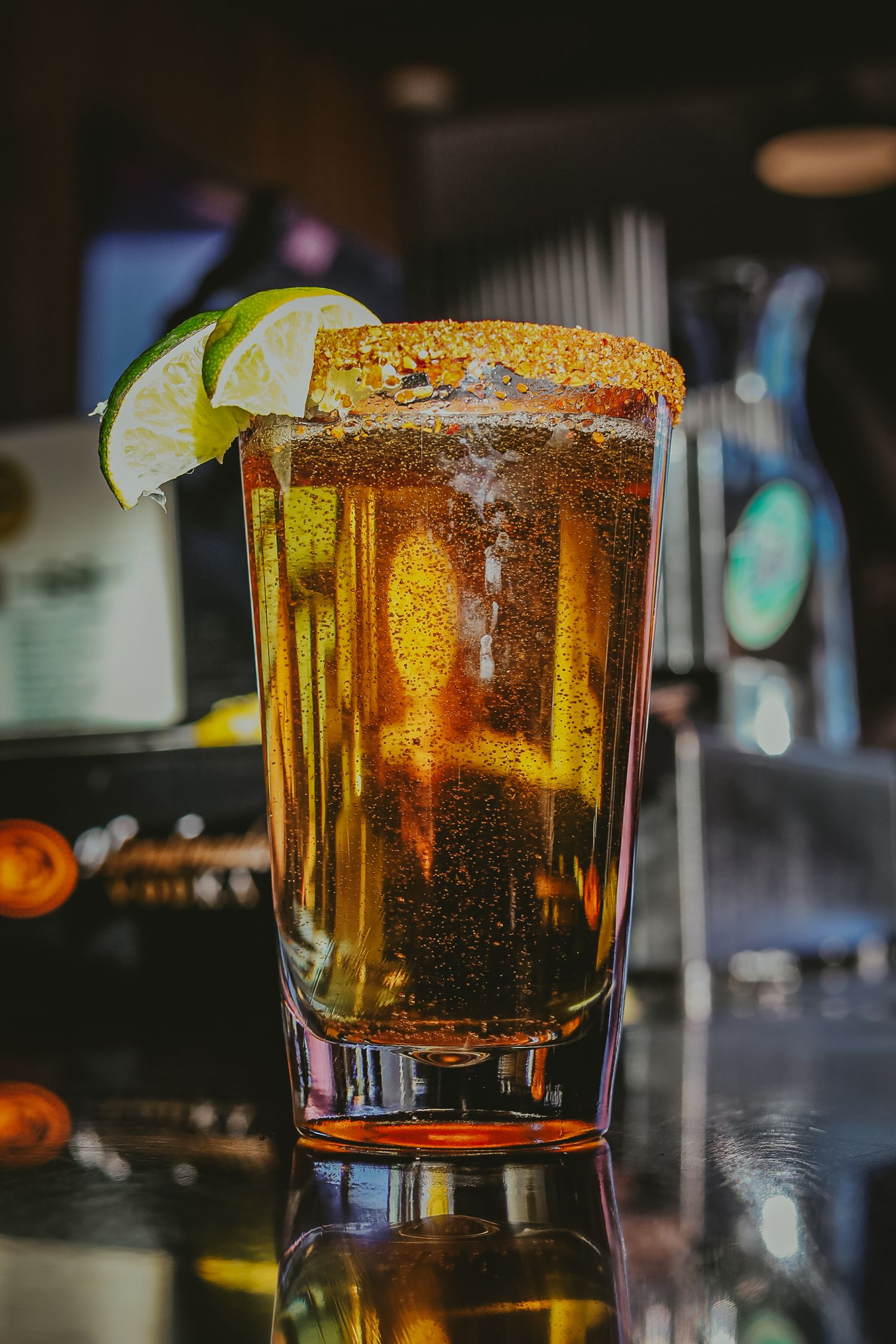 A tall glass of golden beer with a lime garnish and spiced rim sits on a reflective bar counter in moody lighting.
