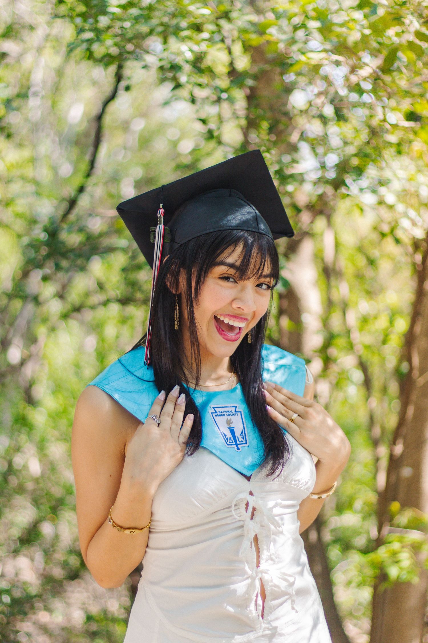 A graduate in cap and gown smiles joyfully while posing outdoors against a sunlit natural backdrop on graduation day.