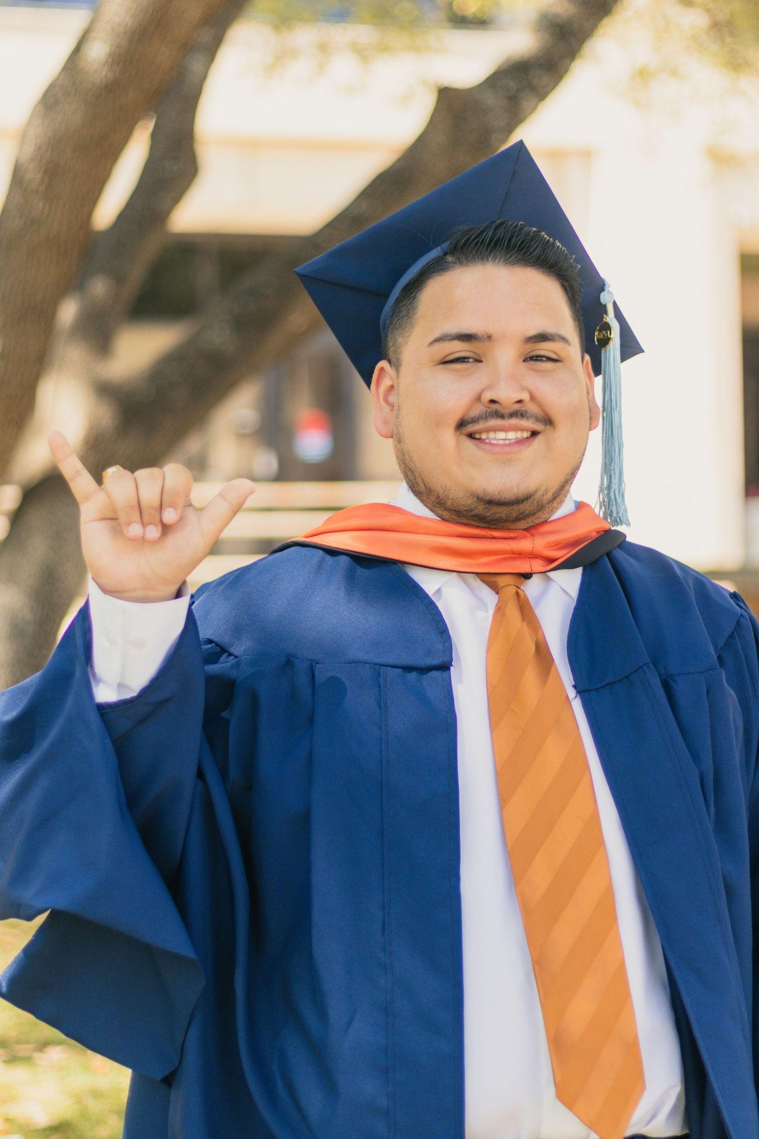 Graduate in blue cap and gown smiles while holding diploma outdoors.
