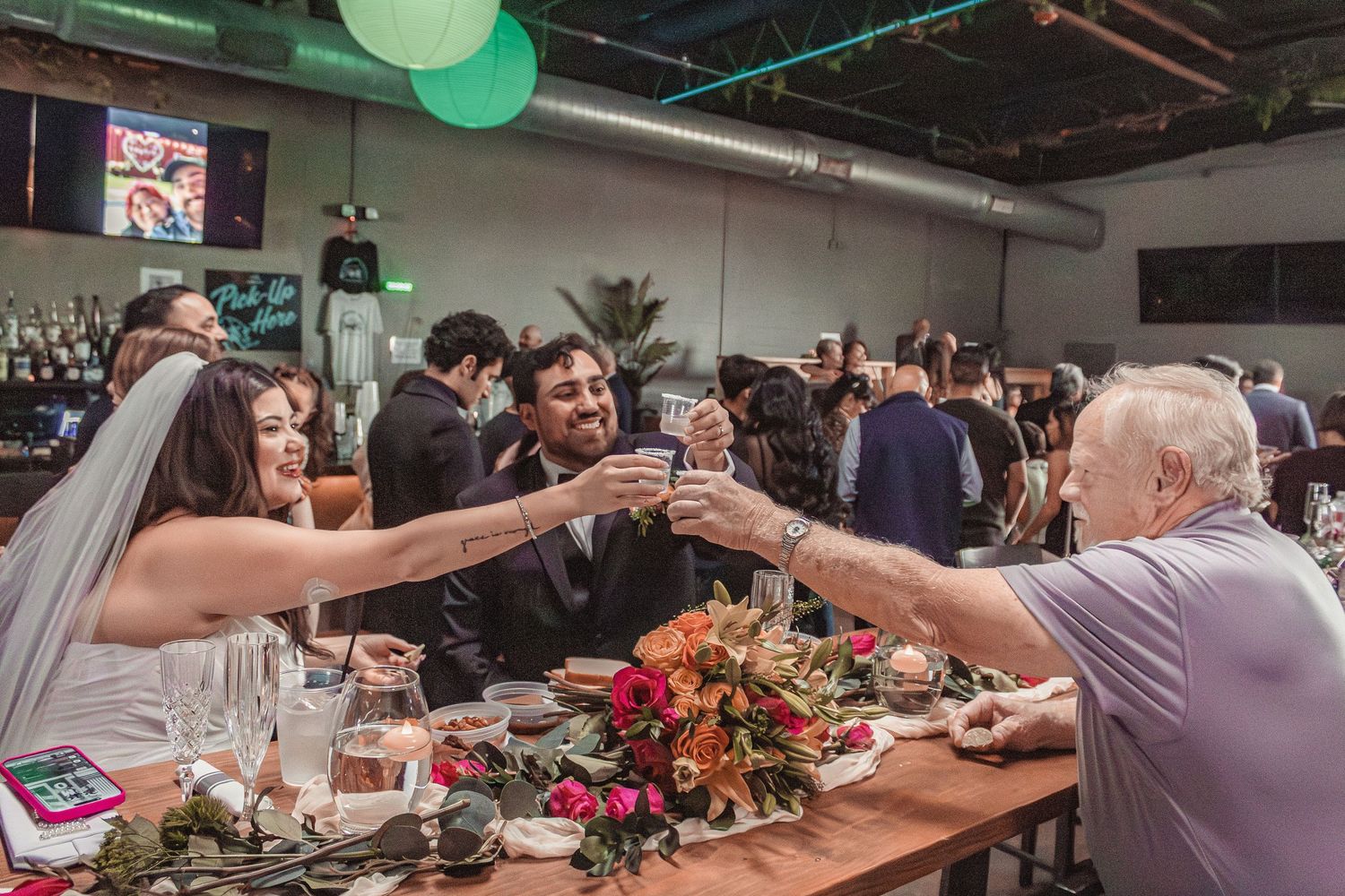 Wedding reception guests toast across a decorated table with flowers at River North Icehouse in San Antonio, Texas.