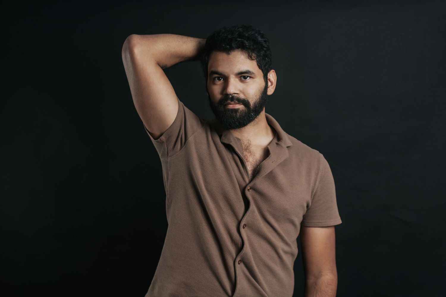 Man in brown polo shirt poses against dark background with hand behind head.