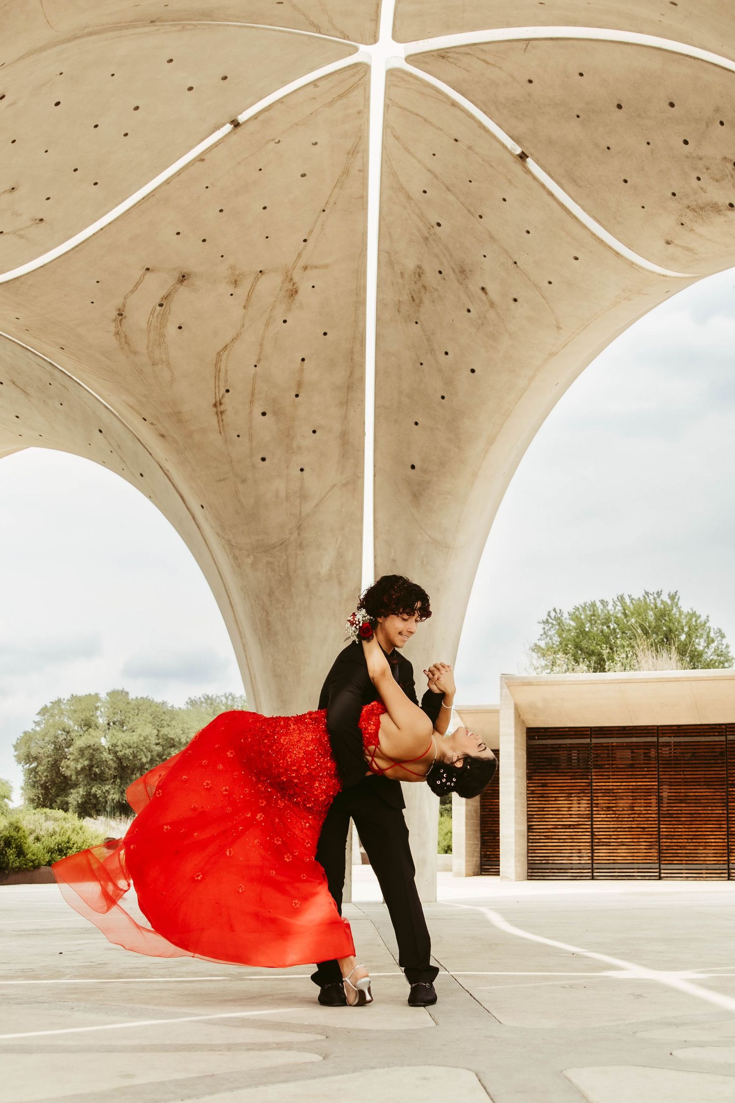 A couple in formal wear dances under a modern architectural structure with flowing red dress movement.