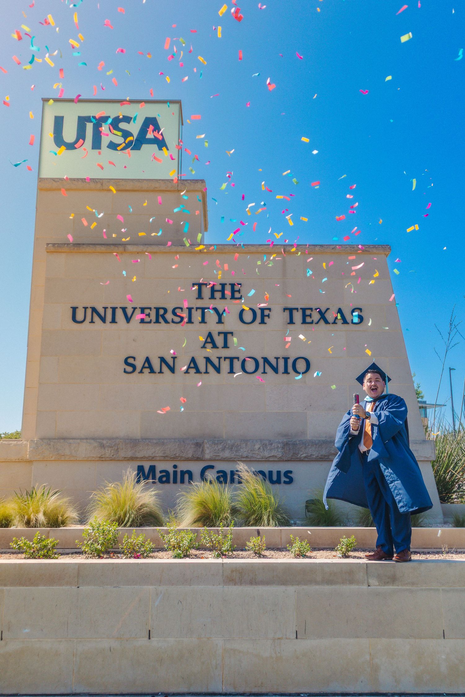 A graduate celebrates at UTSA main campus entrance sign with confetti falling against a bright blue sky.