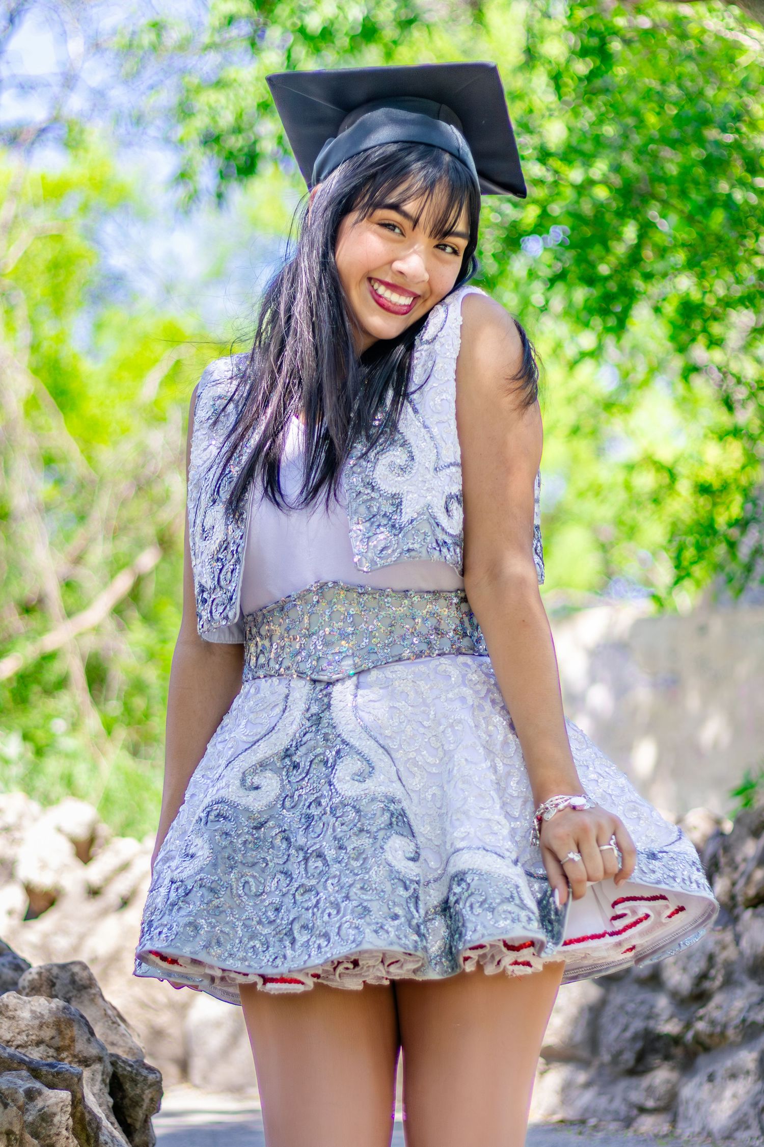 Graduate in silver lace dress and cap poses outdoors among greenery.