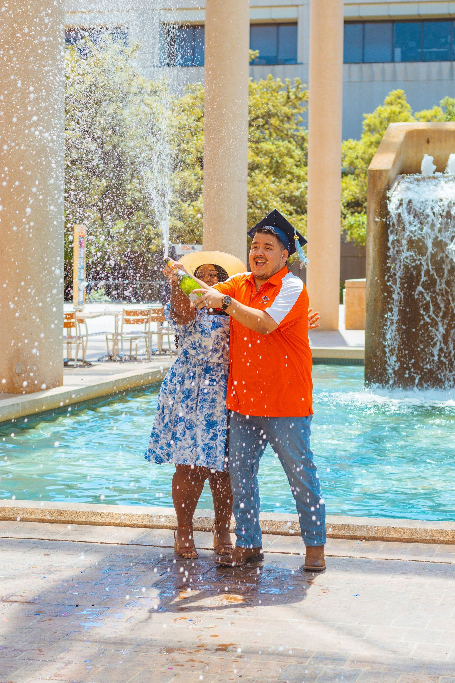 Couple plays joyfully in water fountain spray on sunny day near urban building with fountains in background.