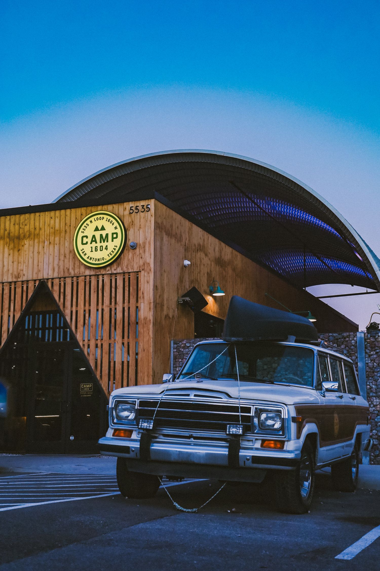 Classic Jeep Wagoneer parked in front of a rustic wooden building with a curved metal roof. Camp 1604 in San Antonio, Texas.