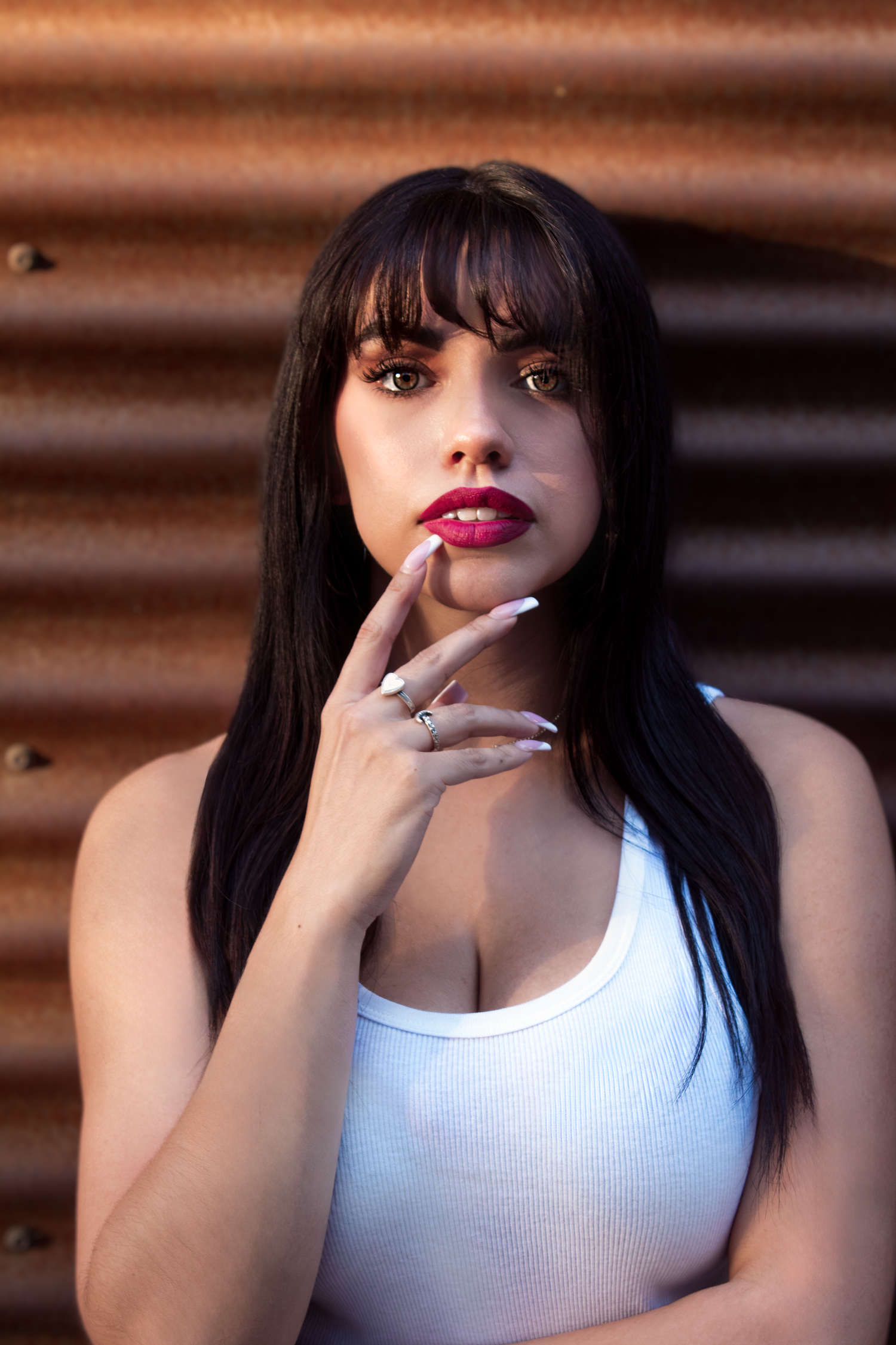 Portrait against a rusty brown wall featuring long dark hair, white tank top, and glossy red lips with metallic rings.