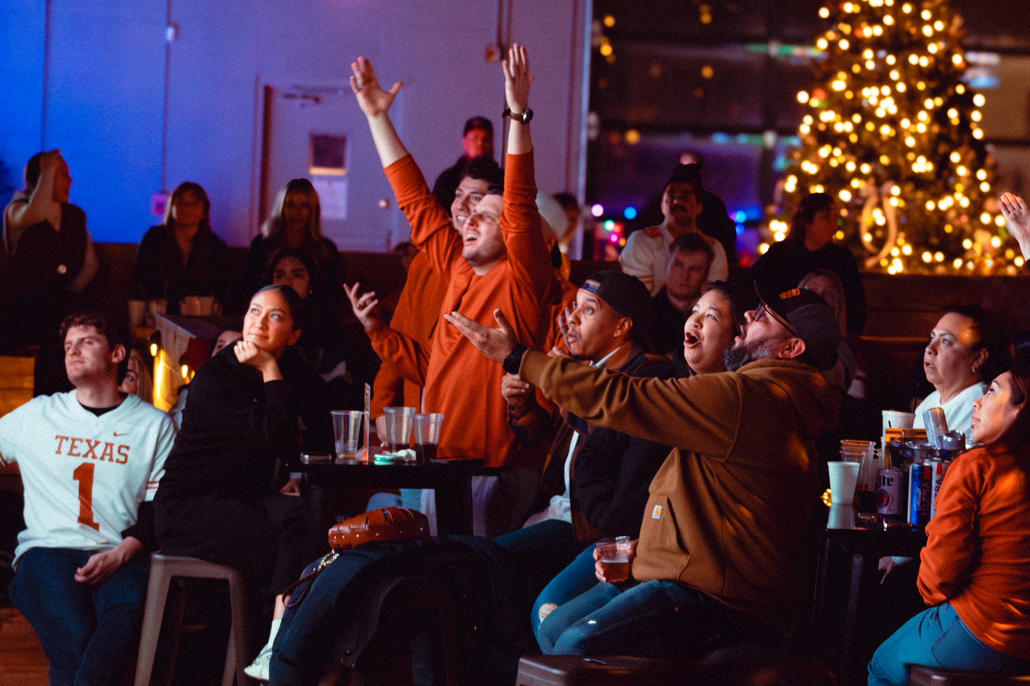 Crowd watching and cheering at an indoor event with Christmas lights in background.