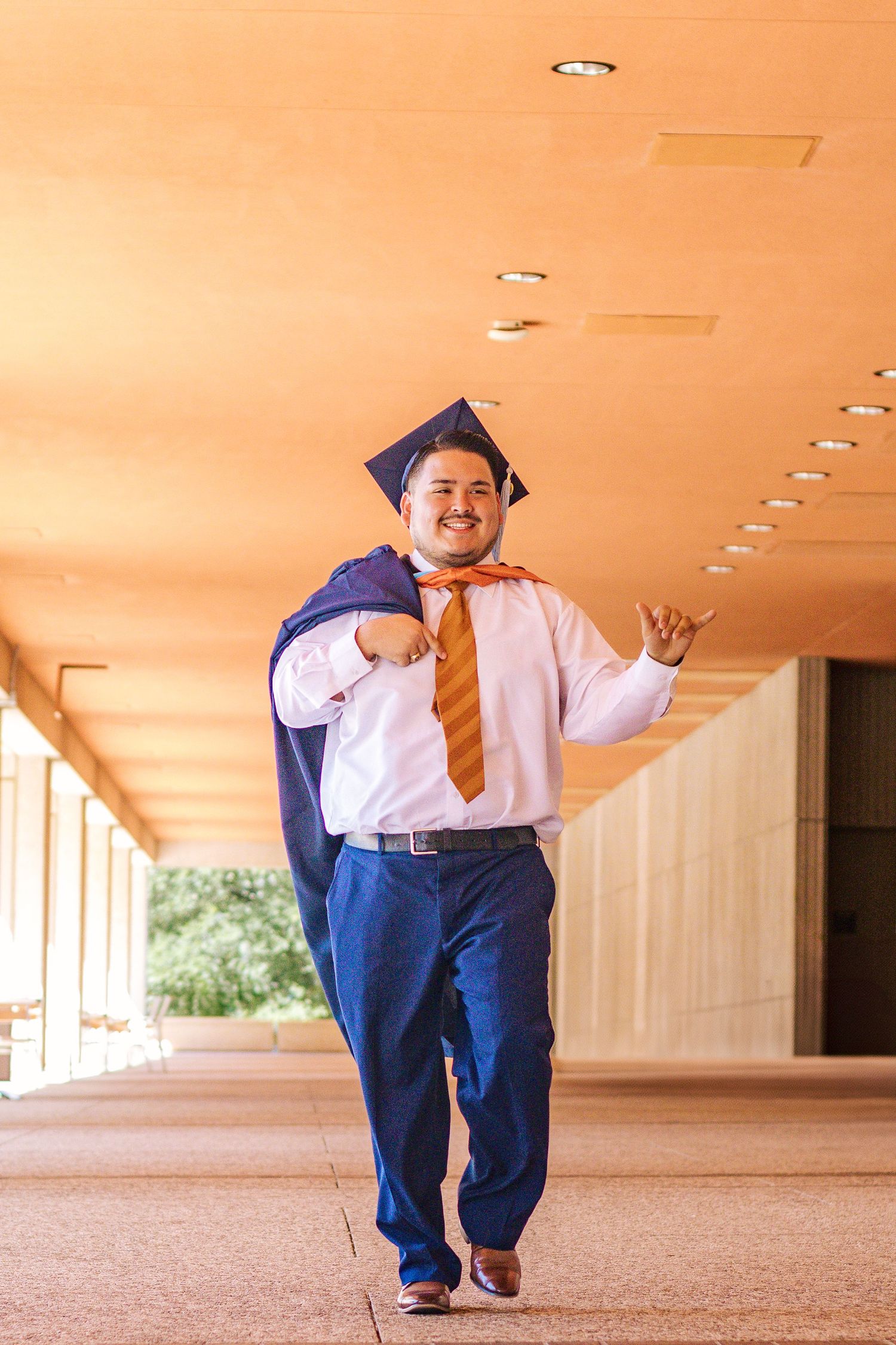 Graduate in blue cap and gown poses cheerfully in hallway with orange lighting.