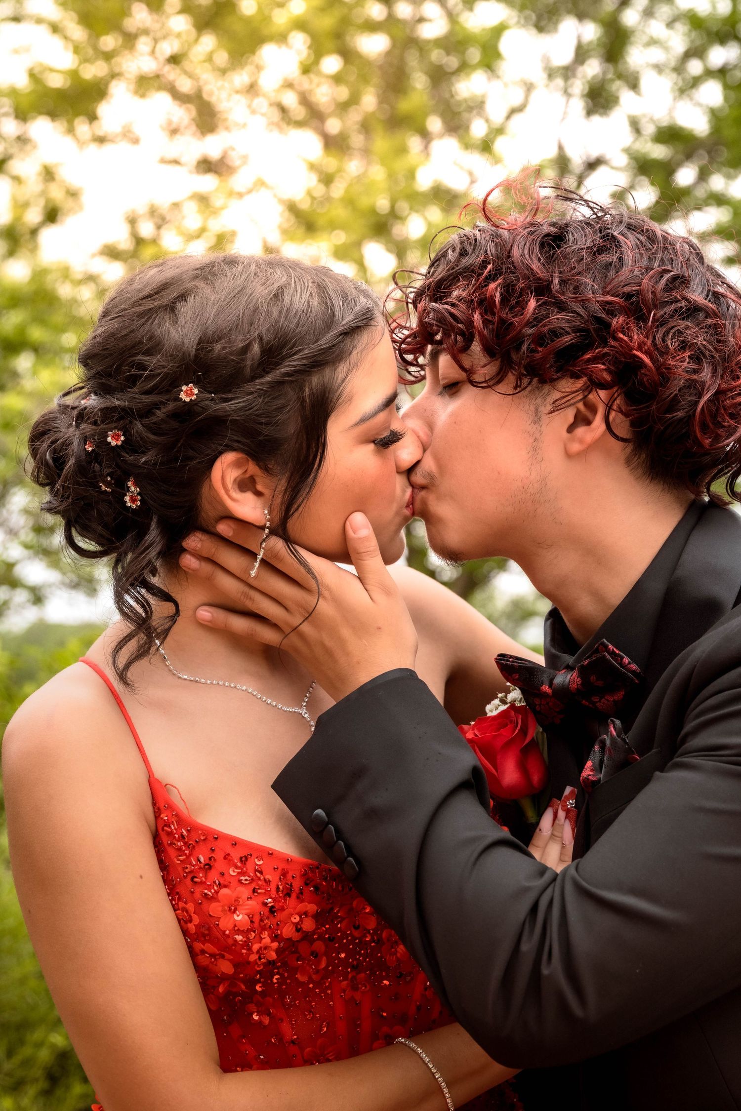 An intimate kiss between a couple in prom attire with a red sequined dress against sunlit trees.