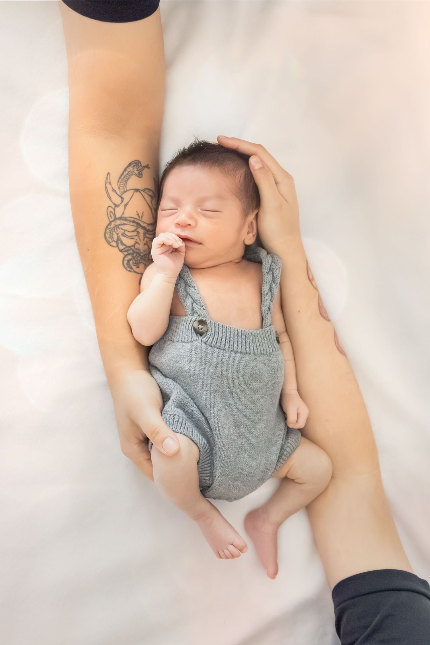 Sleeping newborn in gray knit romper rests peacefully on white blanket as tattooed arm gently touches their head.