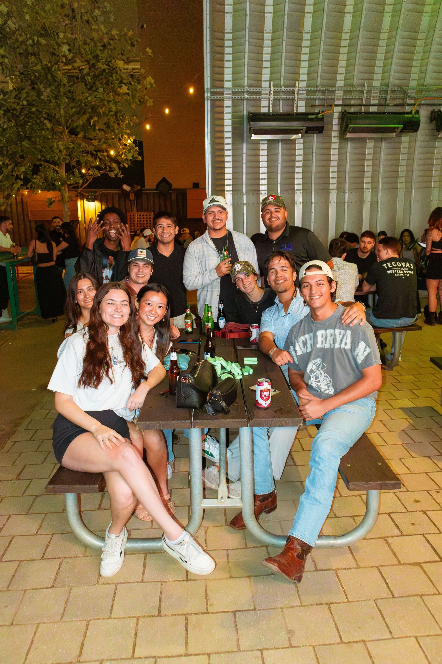Group of friends gathered around a picnic table outdoors enjoying a casual evening together at Camp 1604 in San Antonio, Texas.