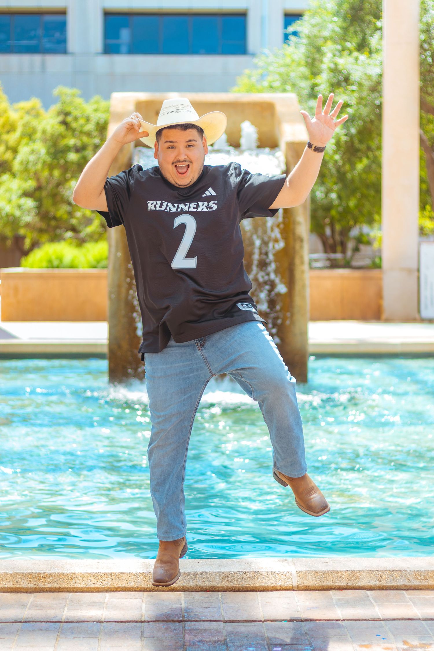 Enthusiastic person in baseball cap and runners shirt jumps playfully by pool fountain.