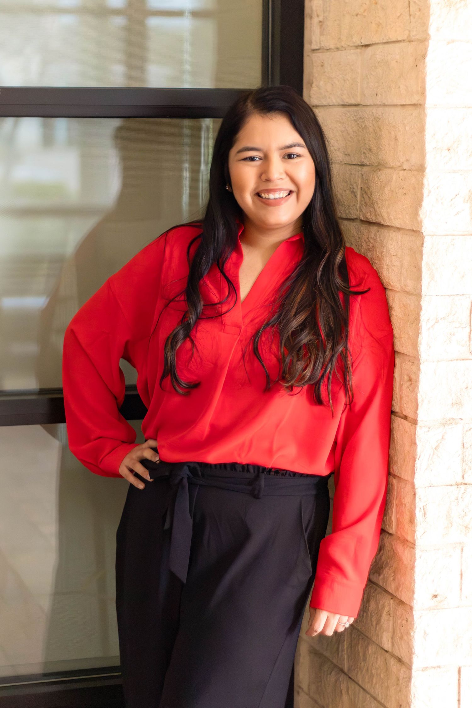 Professional portrait of someone in a bright red blouse and black skirt posing against a brick wall with natural lighting.