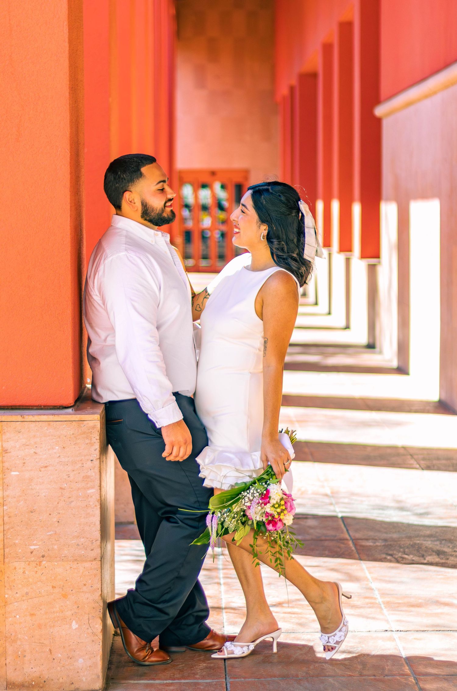 Couple in formal attire posing in coral-colored hallway with floral bouquet.