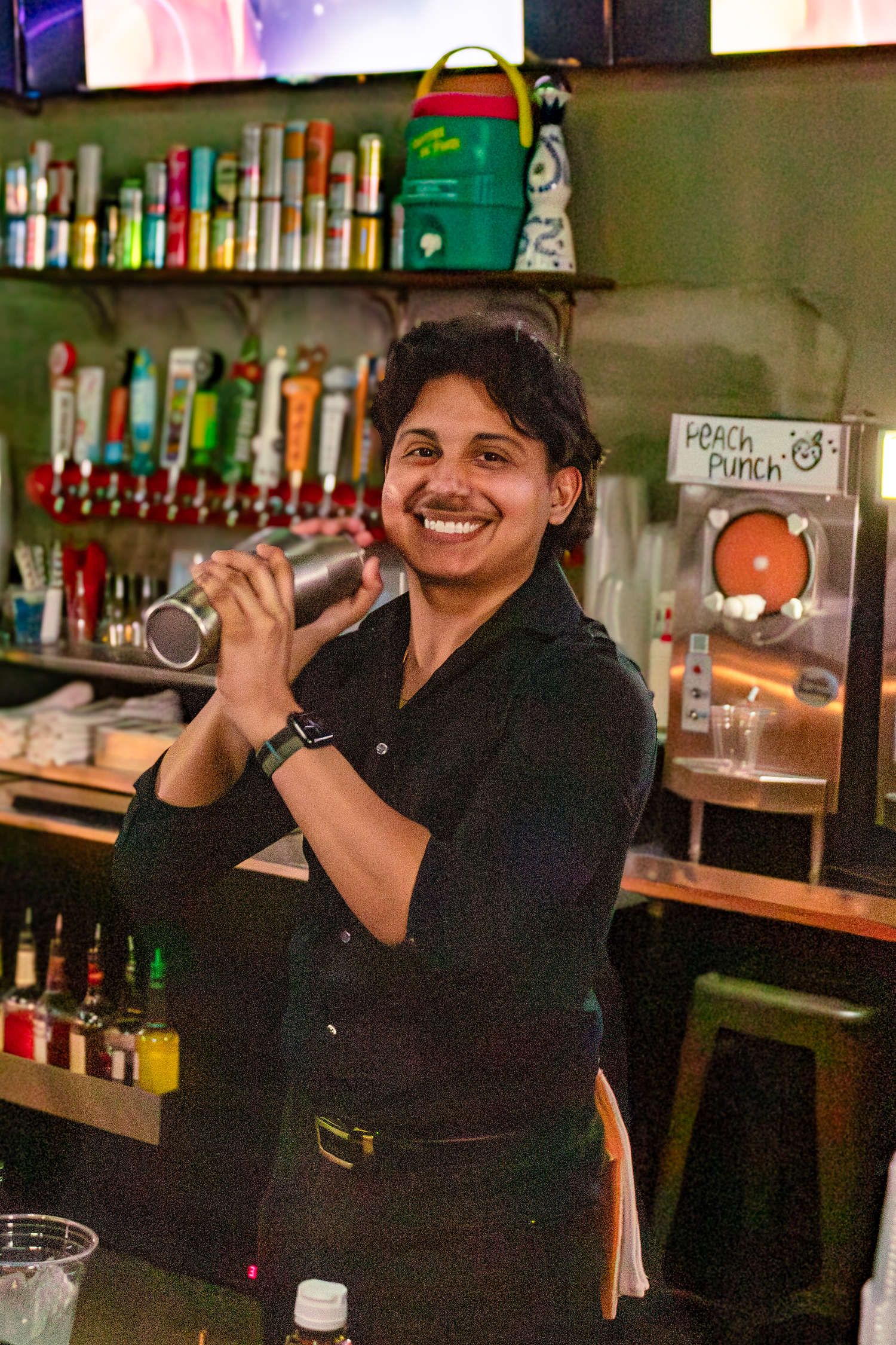 A cheerful bartender poses while working behind the bar with liquor bottles displayed on shelves.